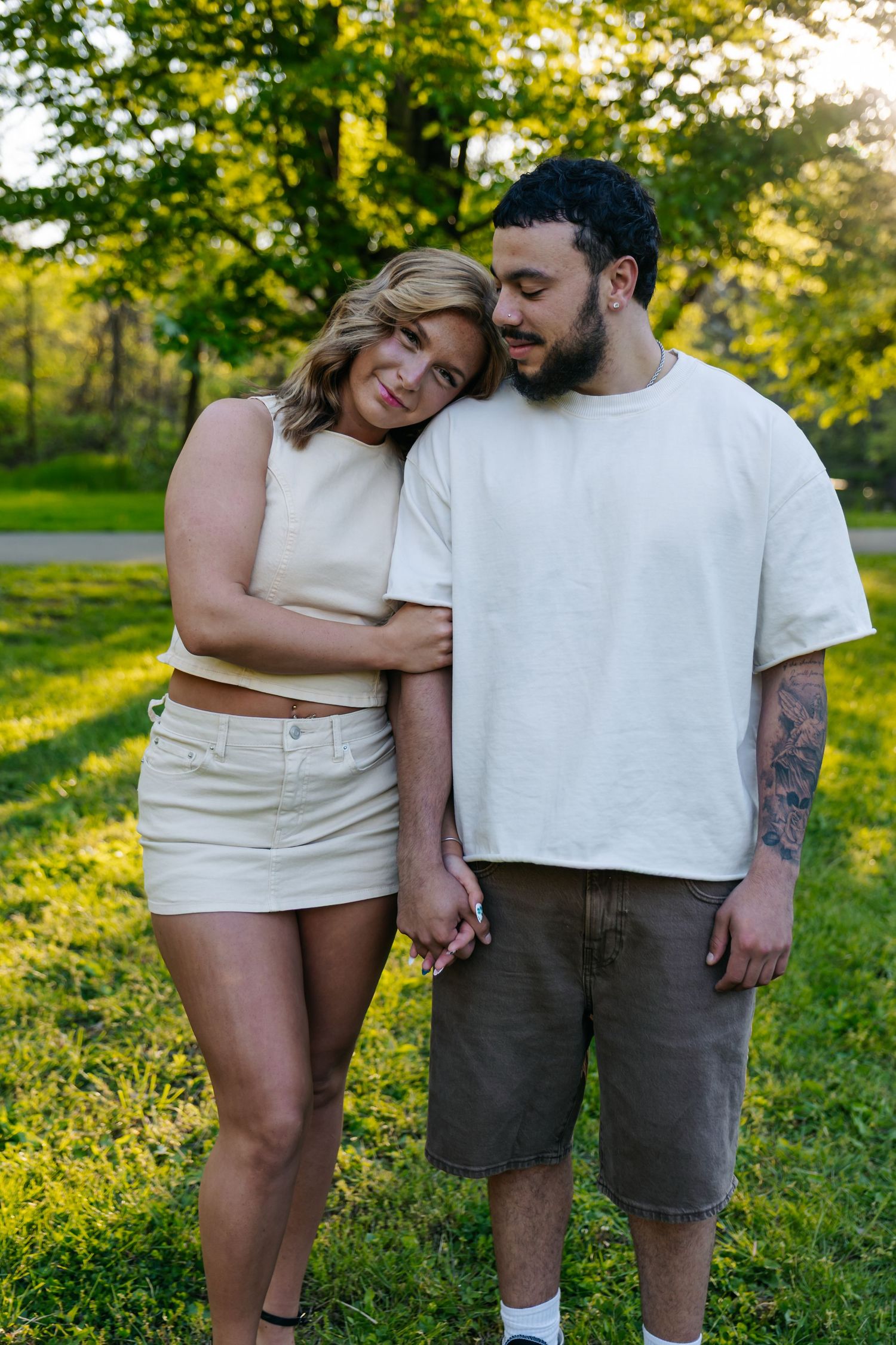A couple holds hands in a sunny park, the woman leaning on the mans shoulder, both wearing casual white outfits.