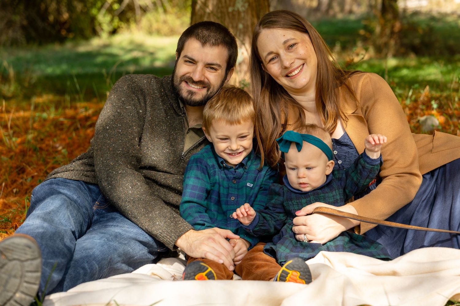 Happy family of four smiling outdoors in autumn, parents with two young children sitting on a blanket among fall foliage.
