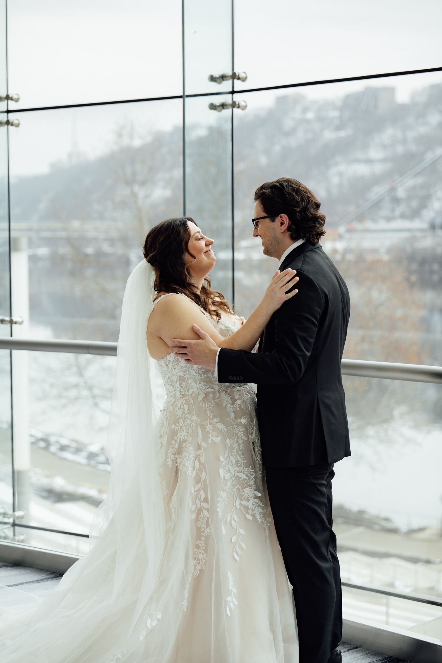 Bride in lace gown and groom in black tuxedo share a romantic embrace by floor-to-ceiling windows on their wedding day.