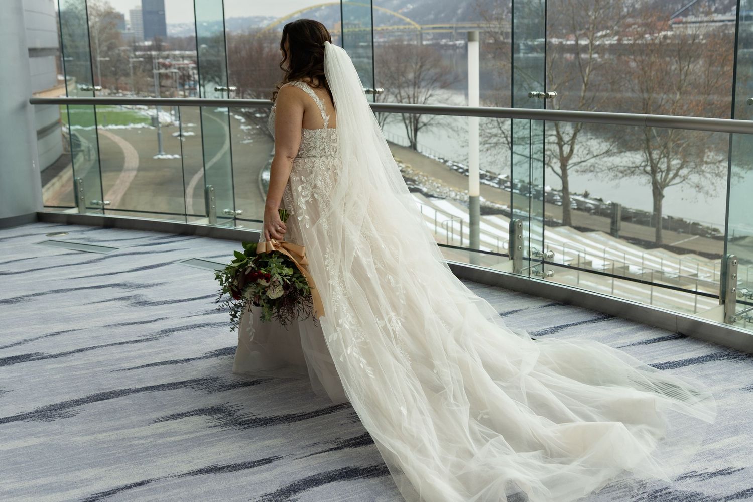 Bride in elegant white gown with long veil holds bouquet on balcony overlooking river.