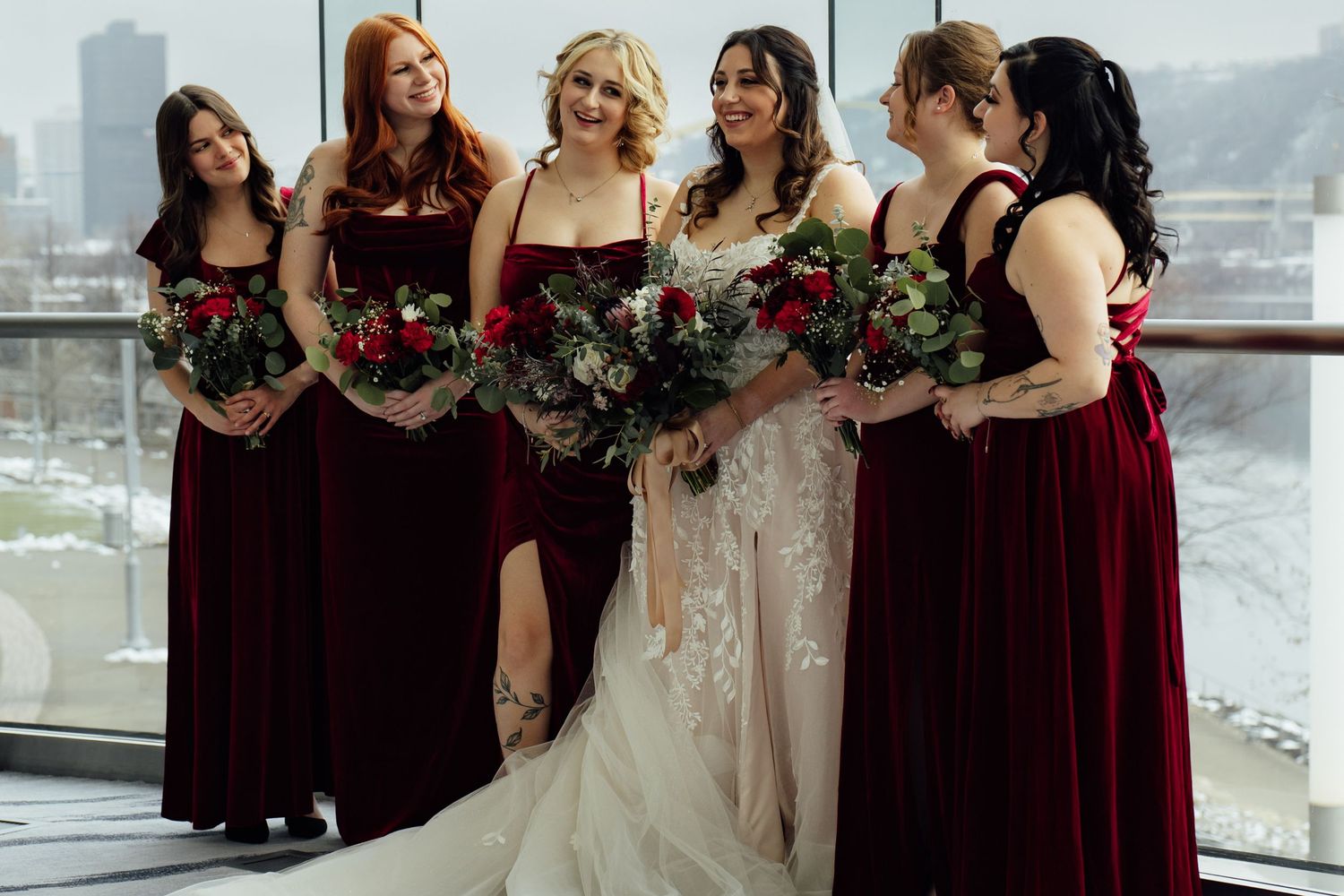 Bride in lace gown with five bridesmaids in burgundy velvet dresses holding red floral bouquets near large windows.