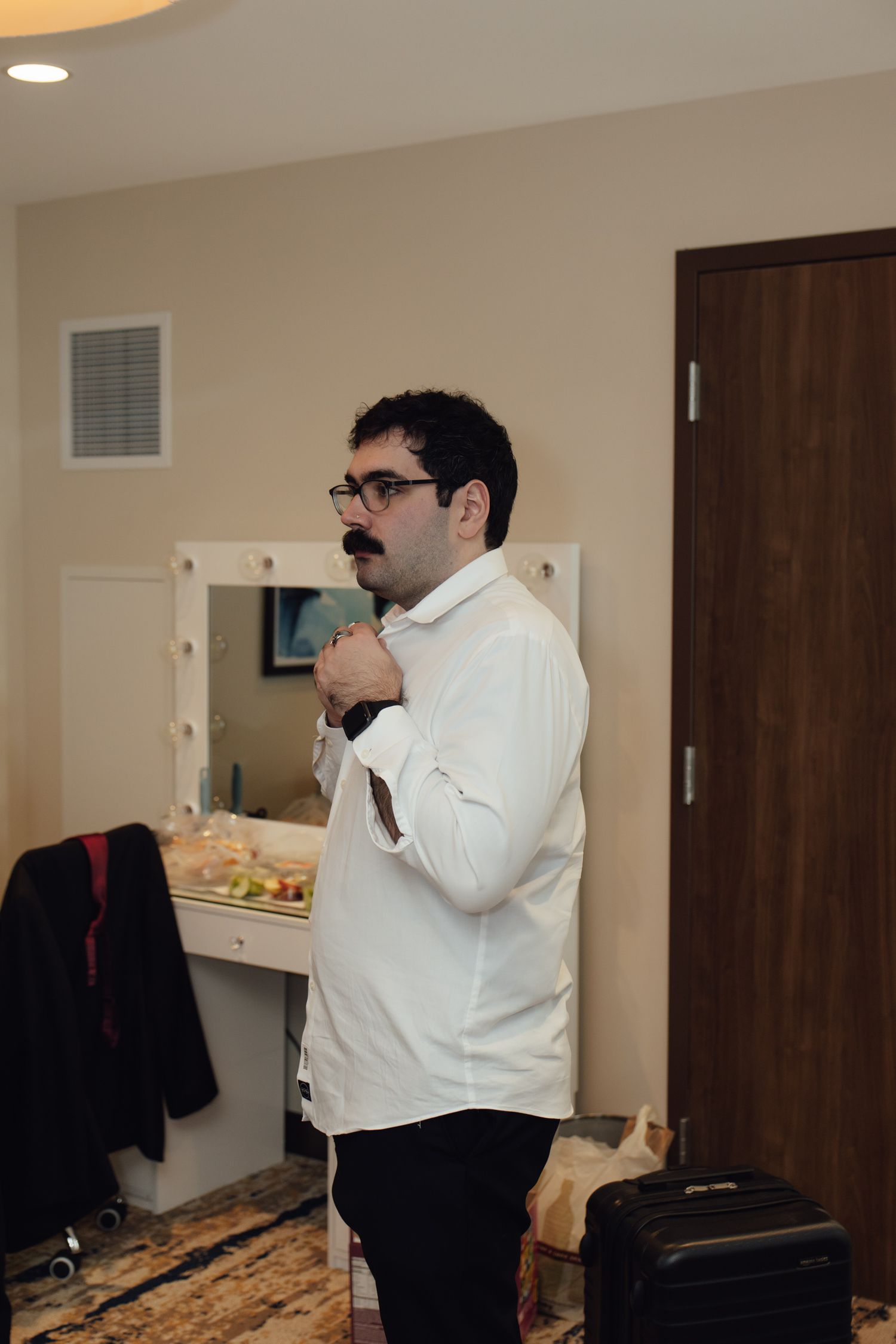 Man in white dress shirt adjusting his tie in a hotel room mirror while getting ready for a formal event.