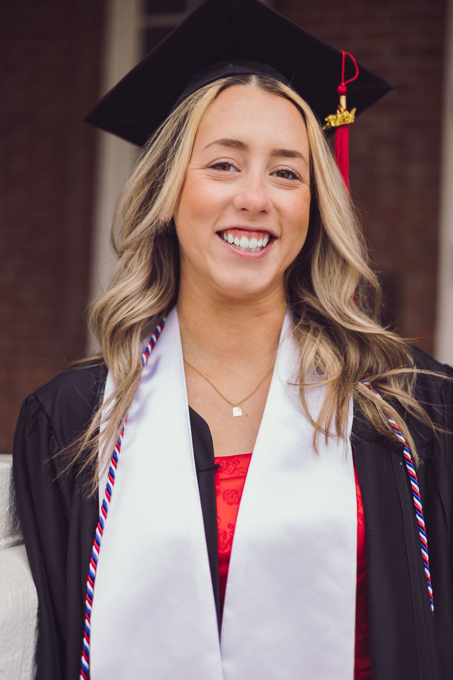 Graduate in cap and gown with white stole smiles at camera in front of brick building.