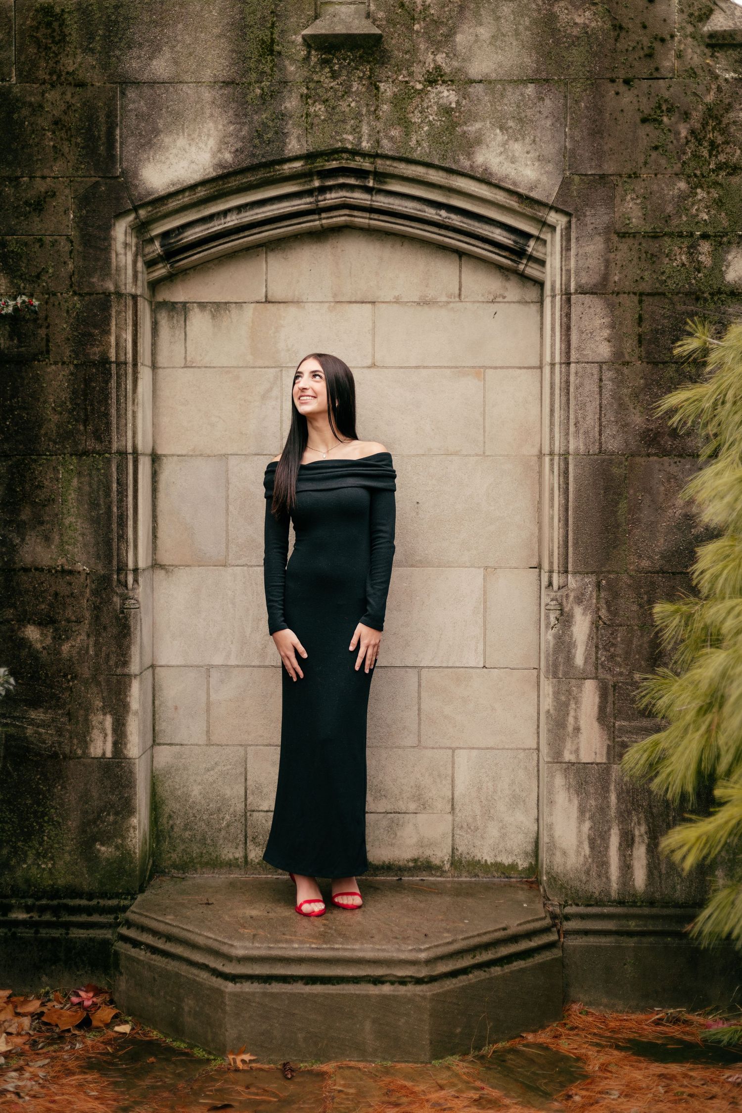 Woman in elegant black off-shoulder dress poses in stone alcove with autumn foliage.