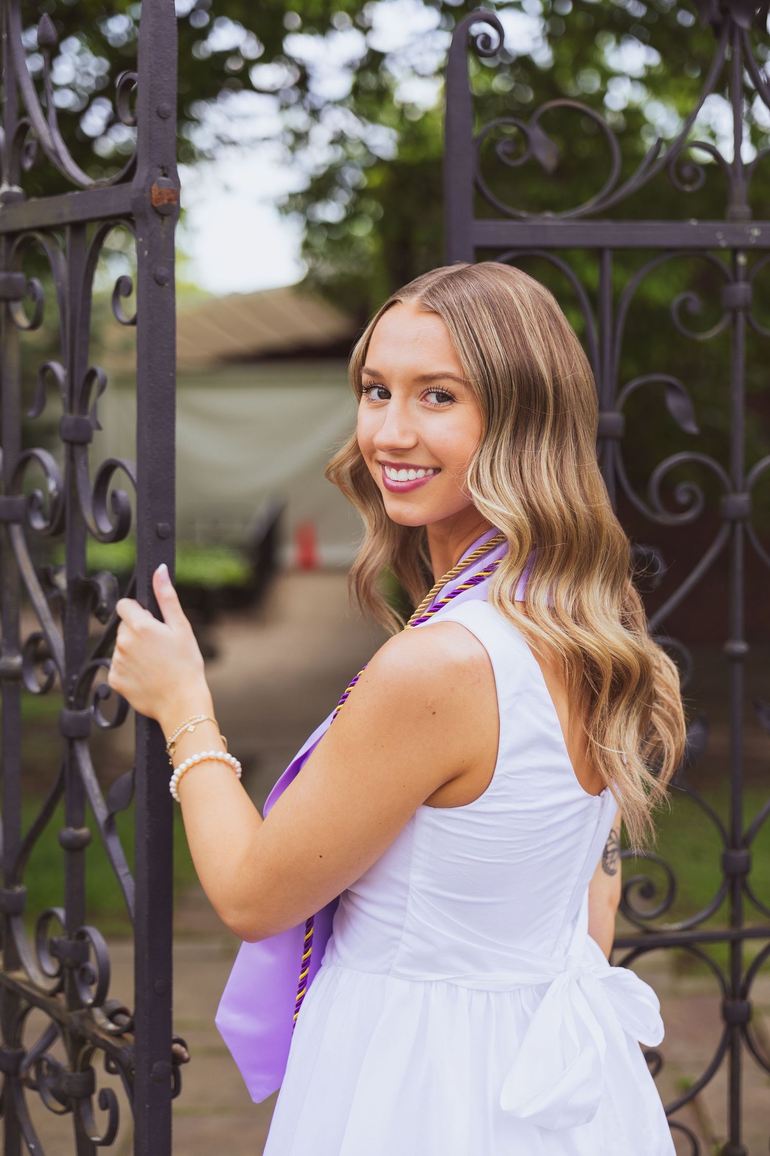 Standing by ornate wrought iron gates wearing a white summer dress and purple backpack with wavy blonde hair.
