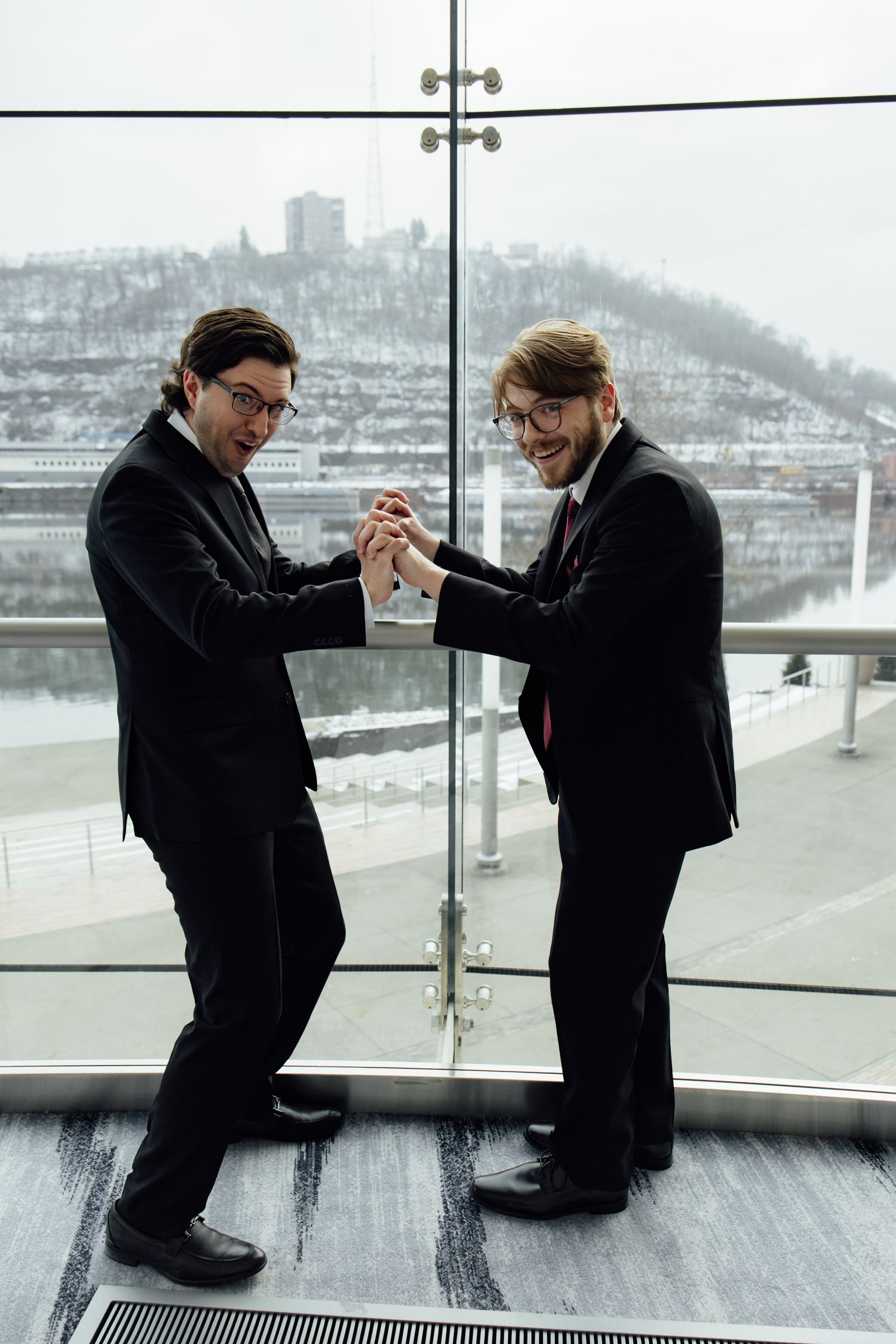 Two businessmen in black suits shaking hands enthusiastically near a glass window with a snowy cityscape backdrop.