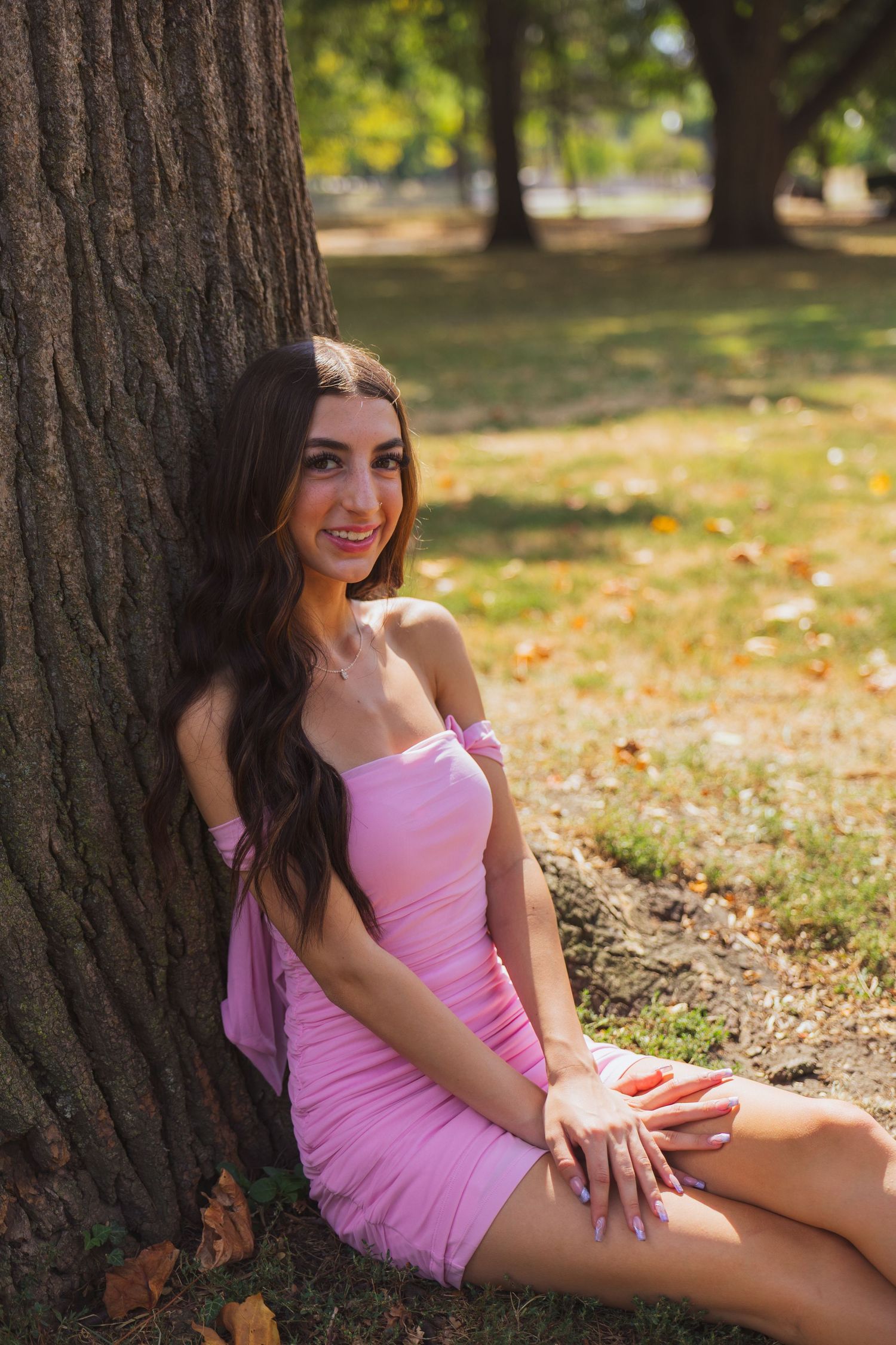 Young woman in pink mini dress sitting against a tree trunk in a sunny park with autumn leaves.