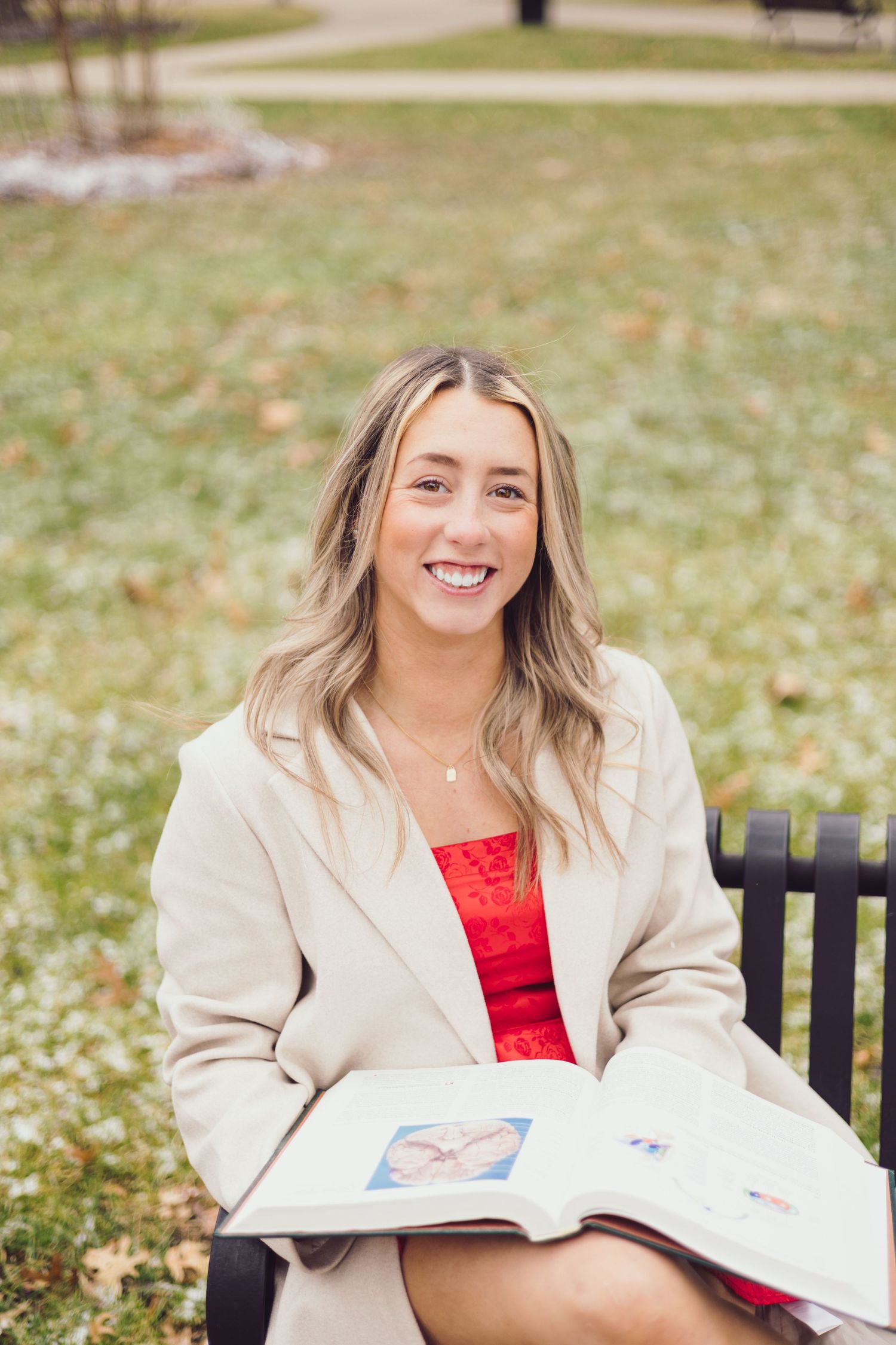 Graduate in cream blazer sits on outdoor bench with open book on college campus.