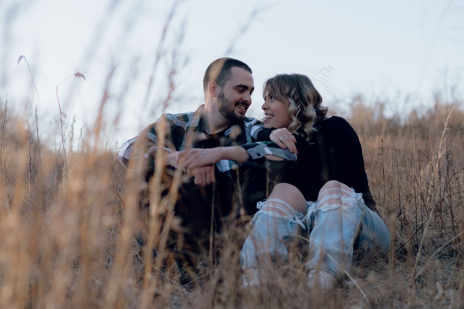 Couple sitting together in tall grass during sunset in rustic outdoor setting.