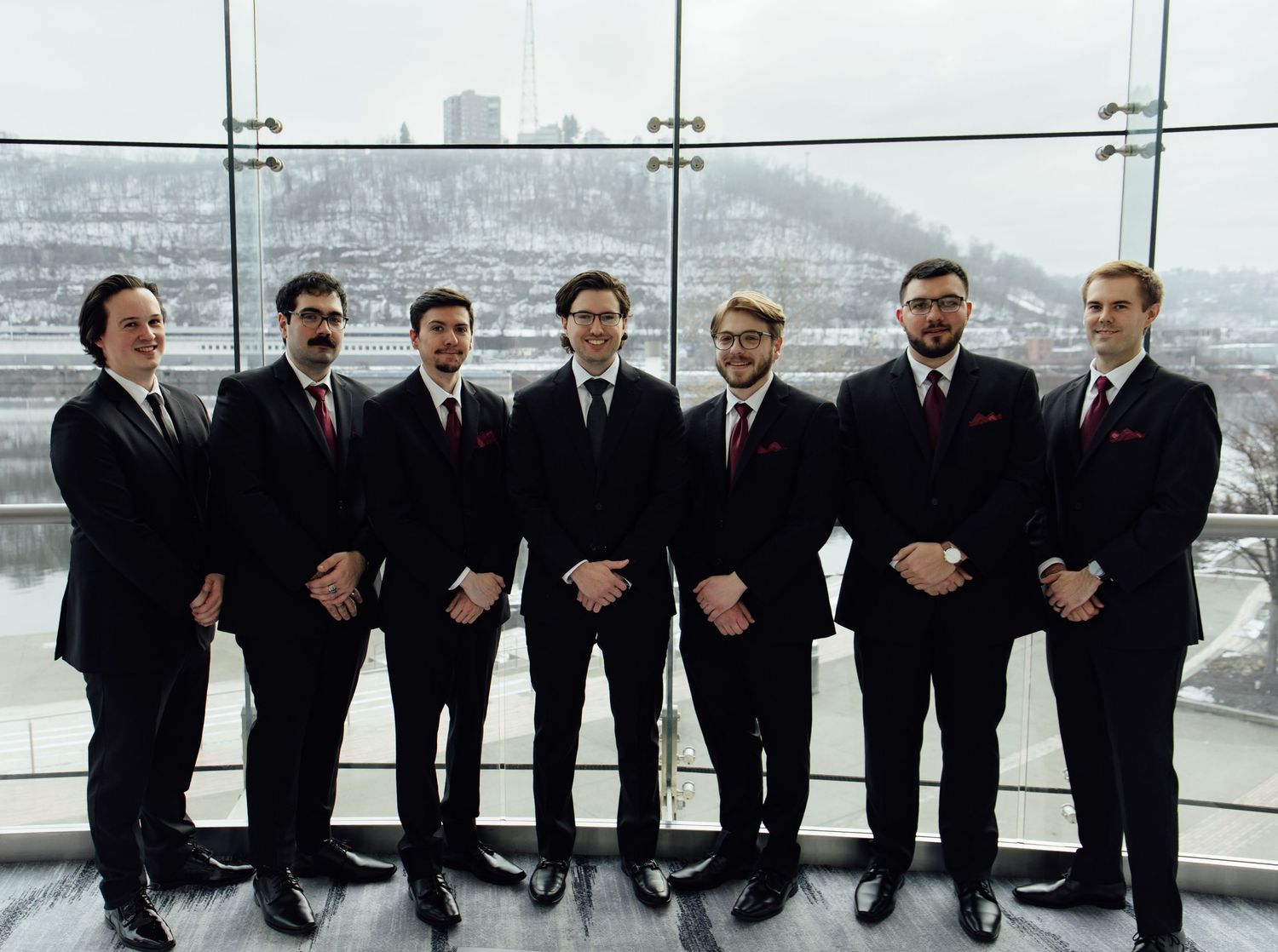Seven groomsmen in black suits with burgundy ties pose formally in a row at a winter wedding venue.