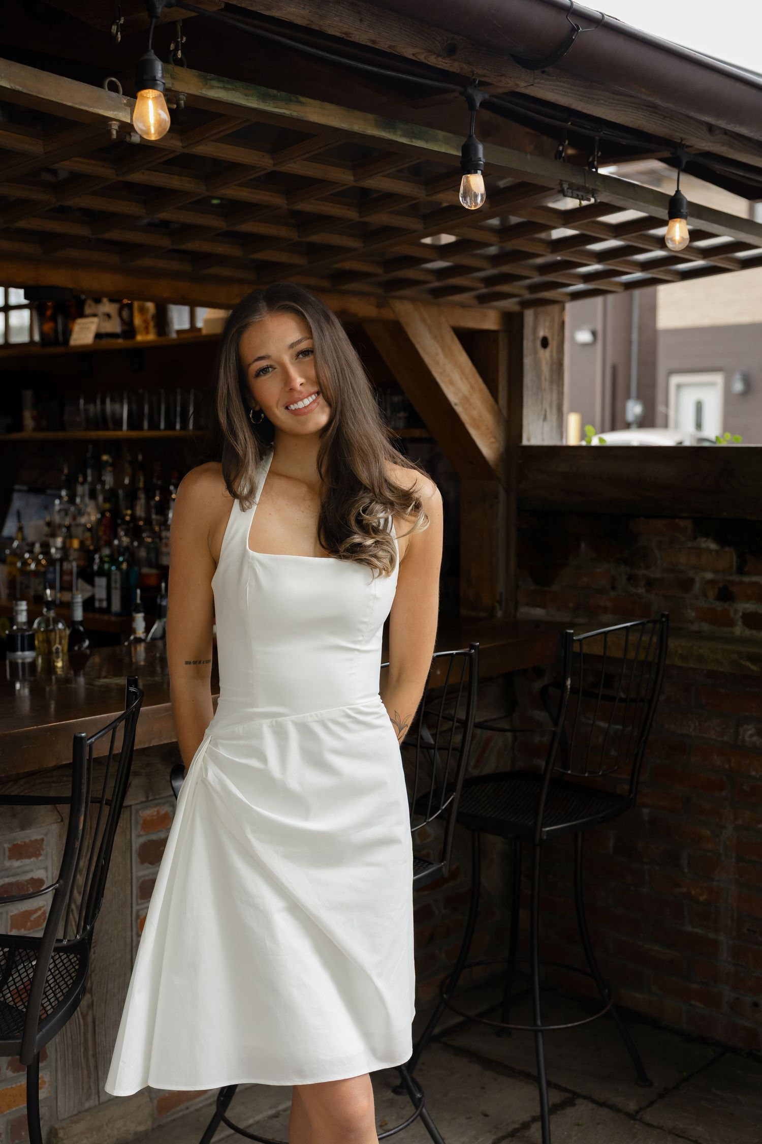 Young woman in white halter mini dress smiling at bar venue with string lights overhead.