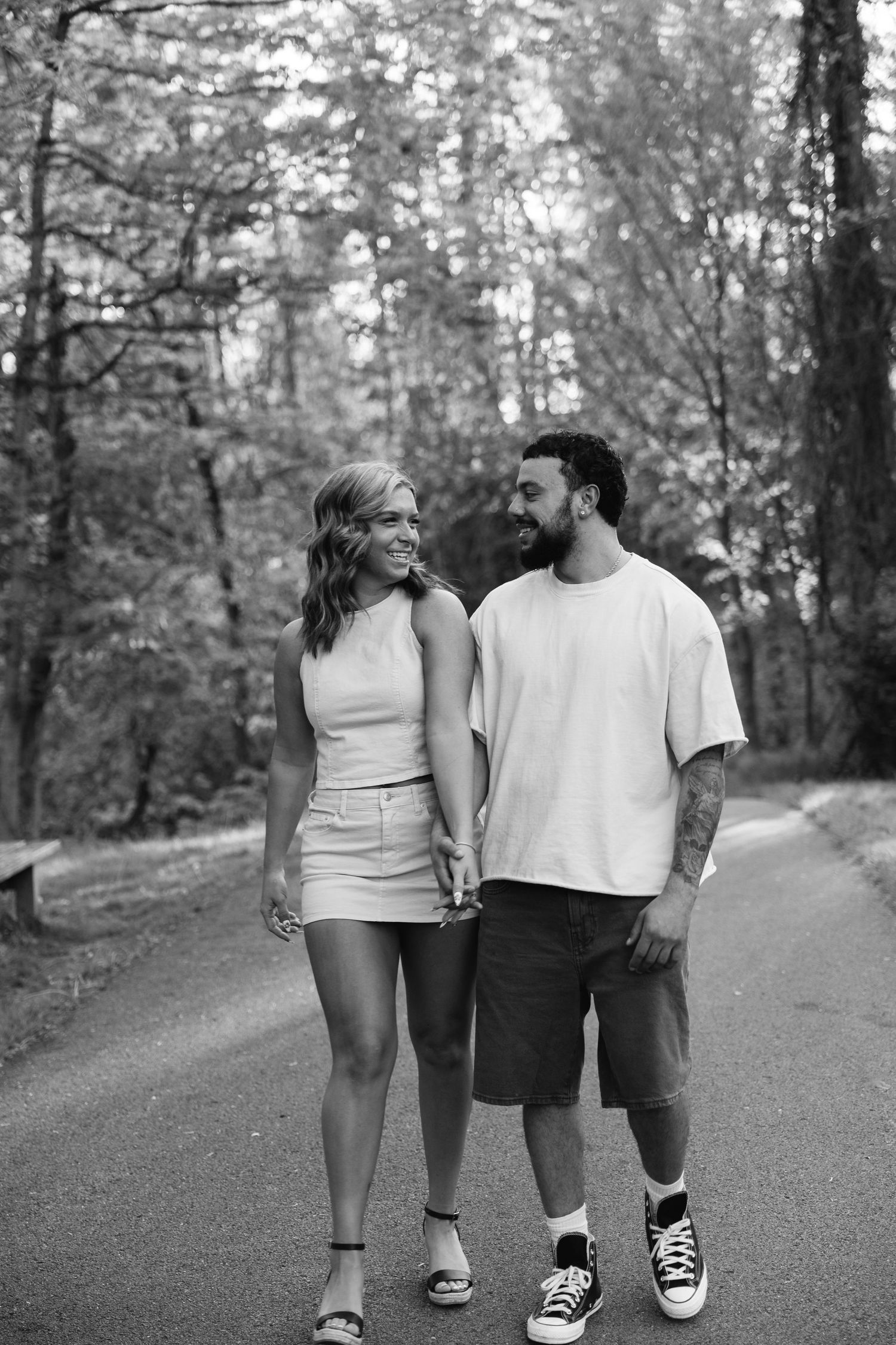 Black and white photo of a happy couple holding hands and walking together on a tree-lined park path.