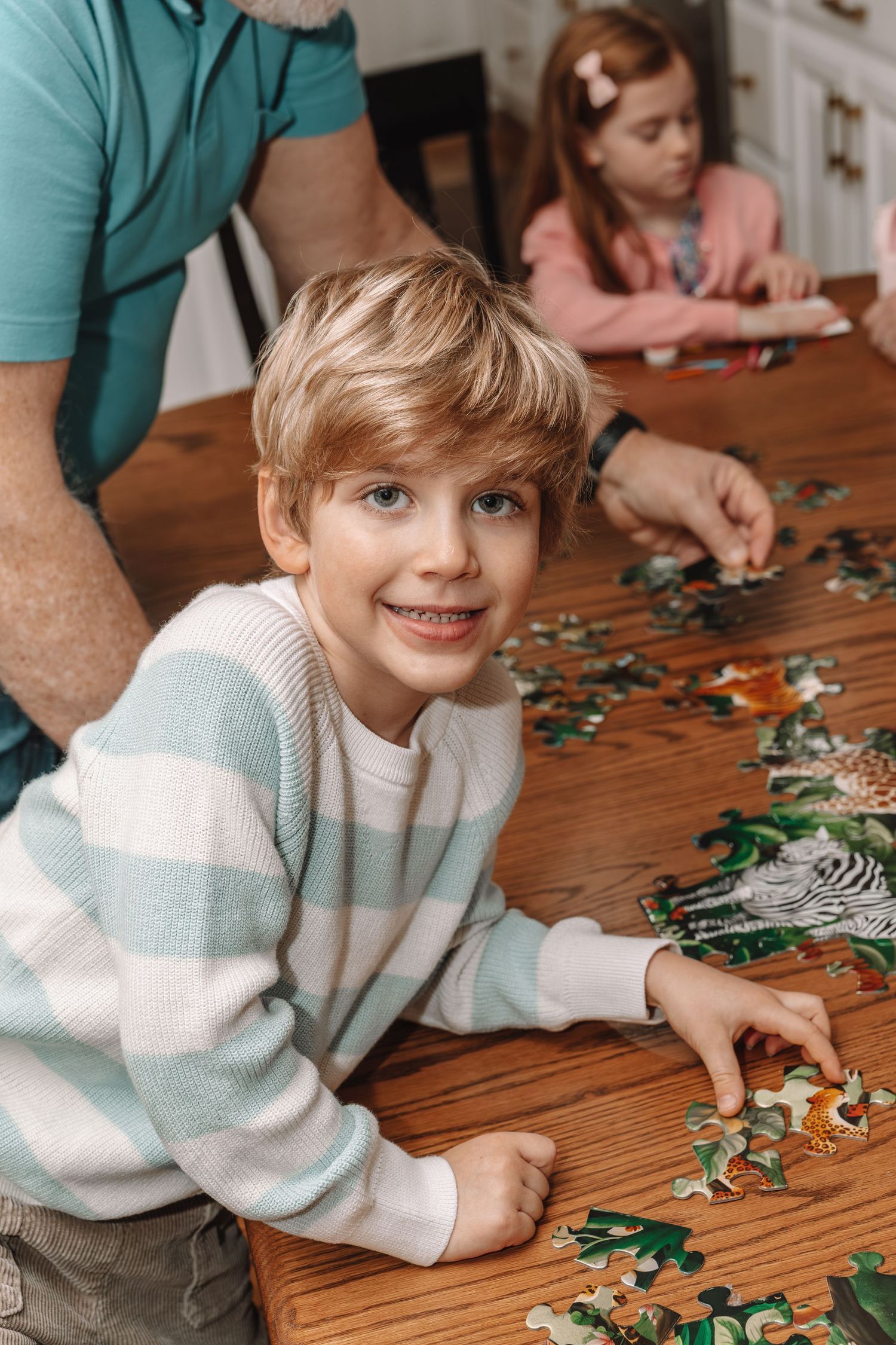 Smiling boy assembles animal jigsaw puzzle at wooden table with father and sister in bright home setting.