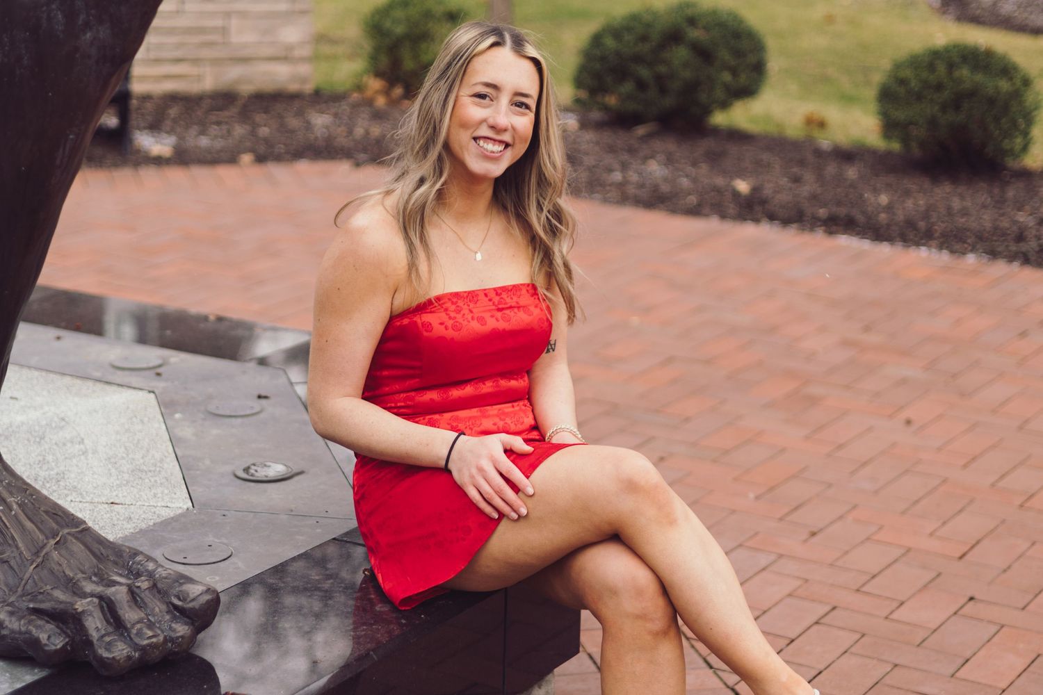 Graduate in red dress sitting and smiling on brick courtyard.