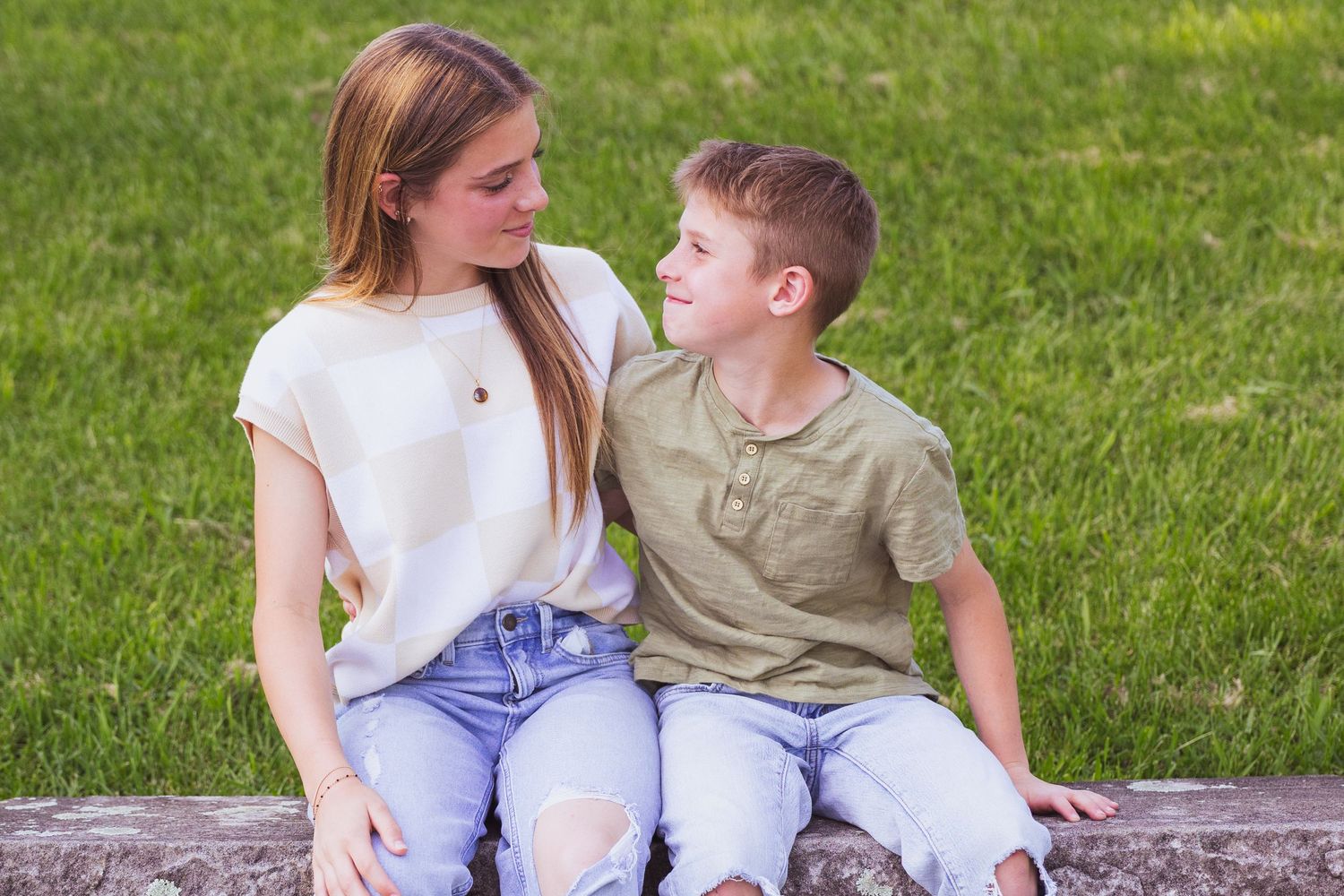 Two young people sitting together on a concrete ledge in a grassy area on a sunny day.