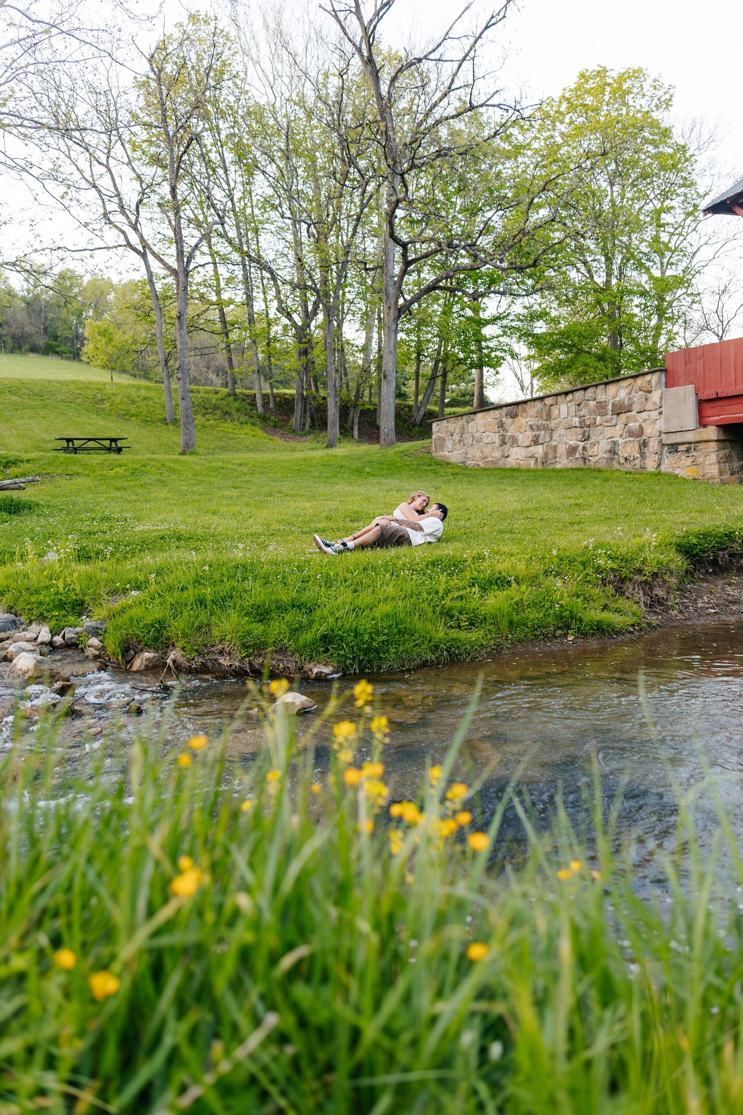 Couple relaxing by a peaceful creek surrounded by lush green grass, wildflowers, and trees in a scenic park setting.