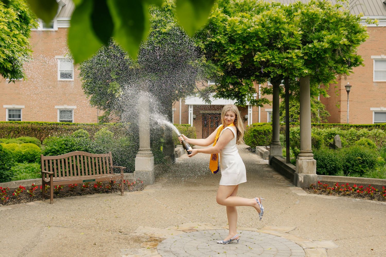 Young woman in white dress joyfully spraying champagne outdoors in a garden courtyard with brick building backdrop.