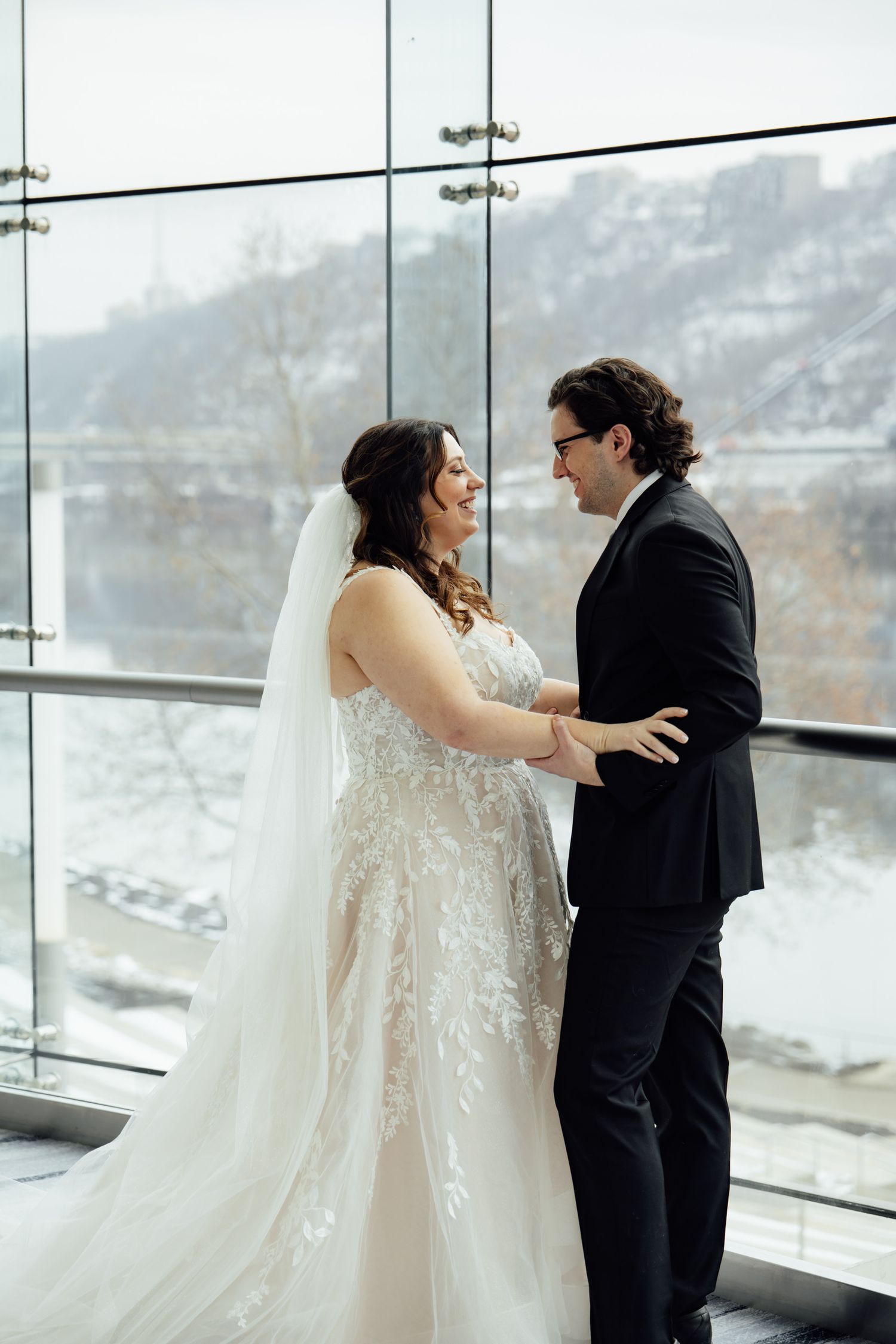 Bride and groom share a romantic moment in front of floor-to-ceiling windows with a snowy winter landscape backdrop.
