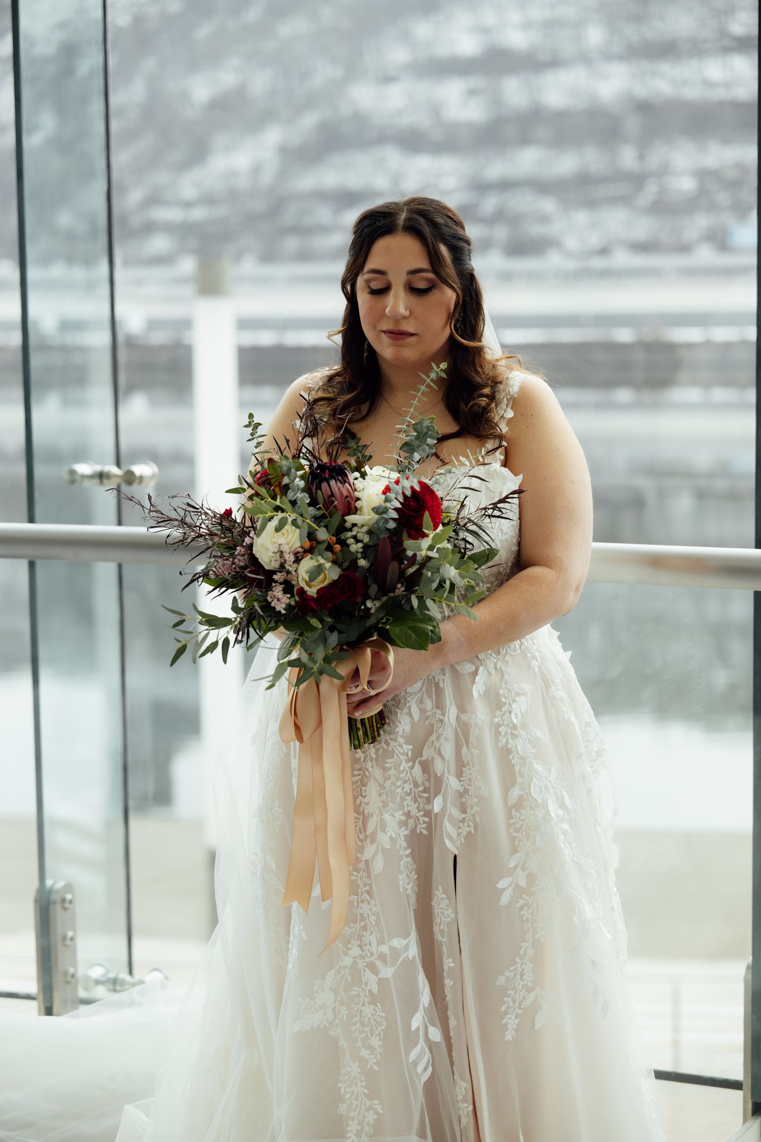 Bride in lace wedding gown holds bouquet of red and white flowers with greenery, standing by glass railing with snowy backdrop.