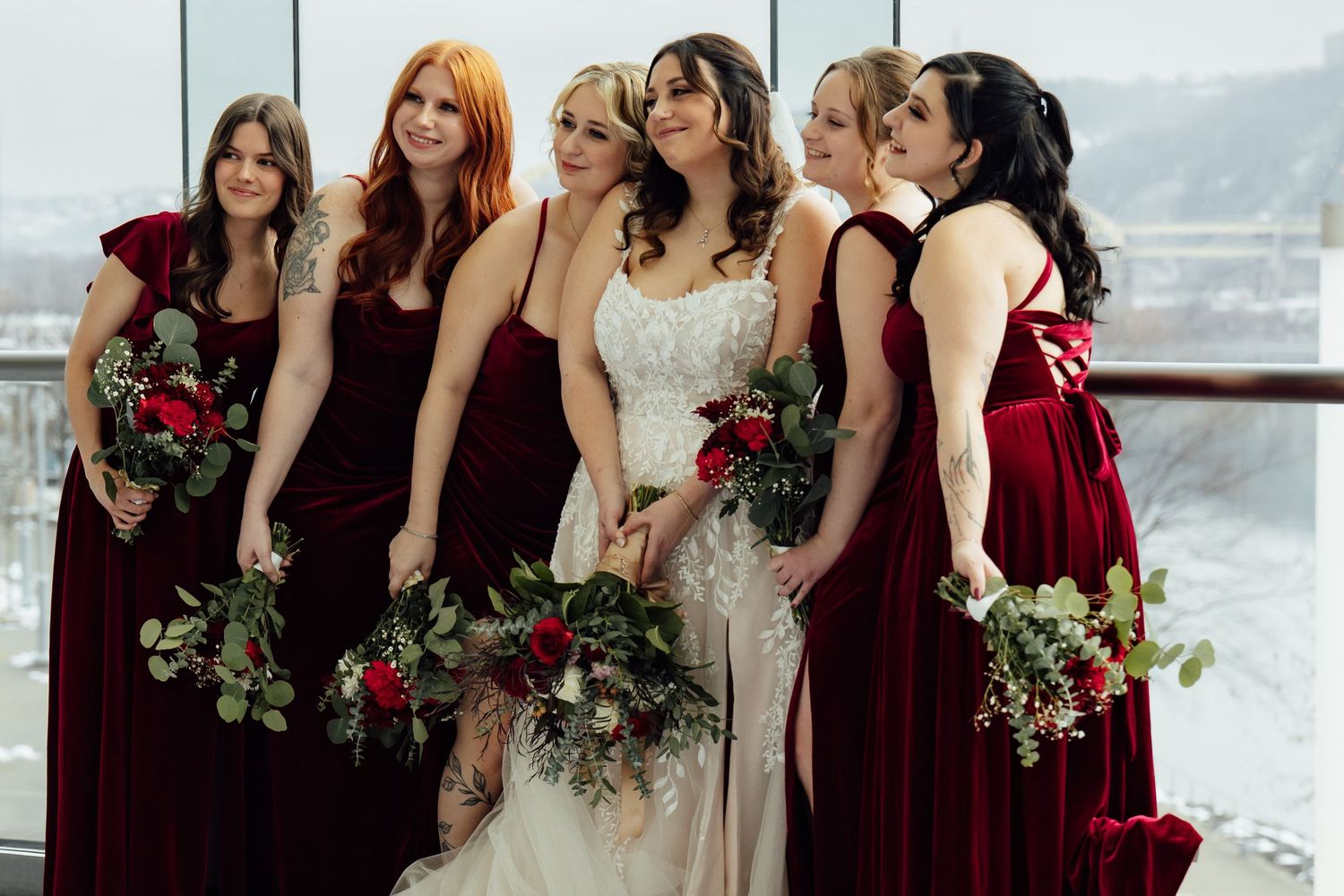 Bride in lace gown with bridesmaids in burgundy velvet dresses holding red floral bouquets near large windows.