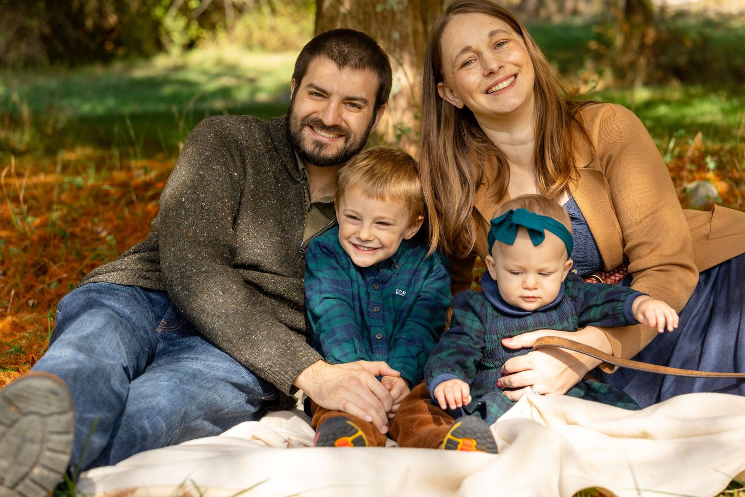 Happy family of four sitting on a blanket outdoors in autumn, parents smiling with toddler son and baby daughter.