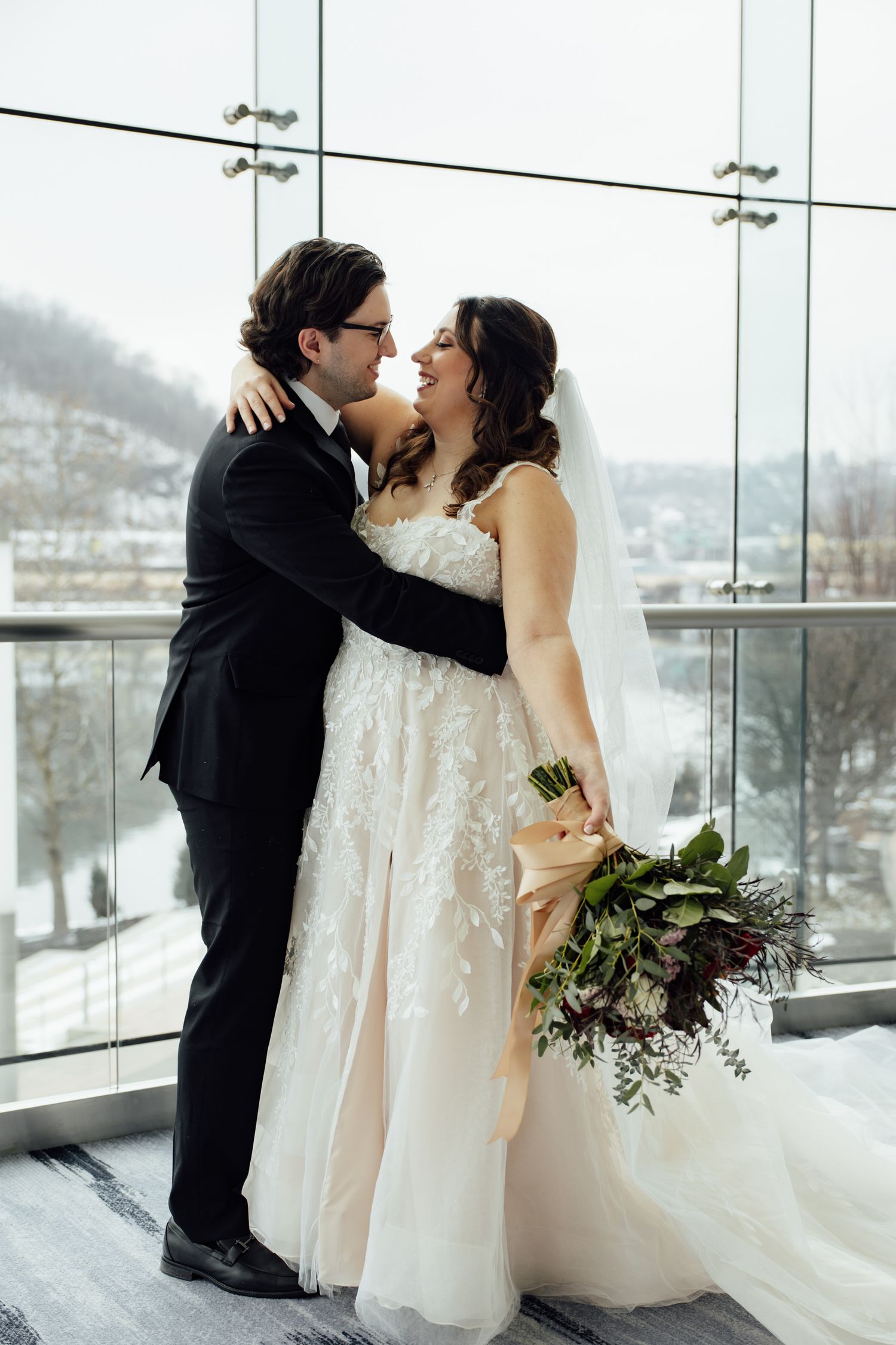Bride and groom share a romantic moment in front of a snowy winter window, bride holding a lush bouquet.