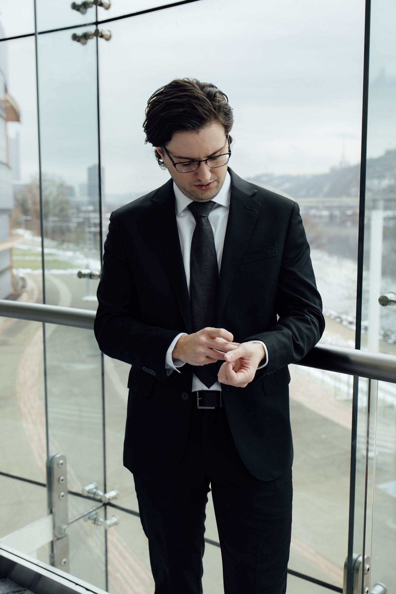 Stylish man in black suit and glasses standing on a glass balcony adjusting his cufflinks.
