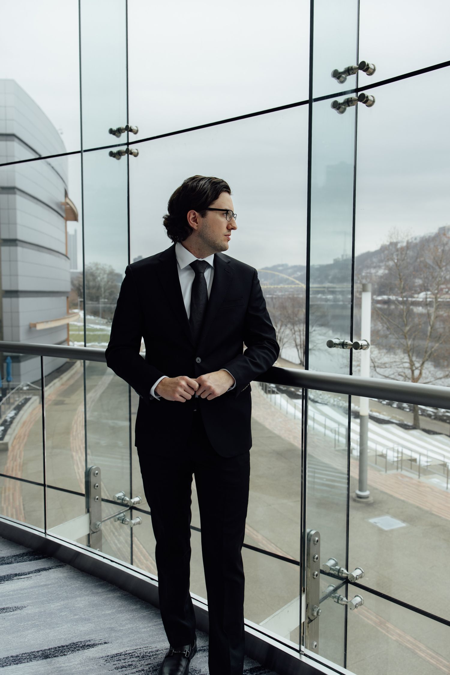 Confident man in black suit standing on glass-walled corporate building balcony.