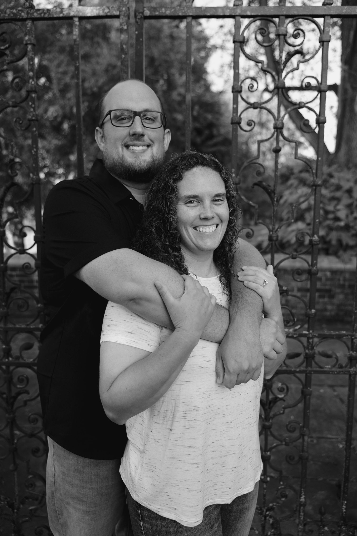 Black and white photo of a smiling couple embracing in front of an ornate iron gate surrounded by lush greenery.