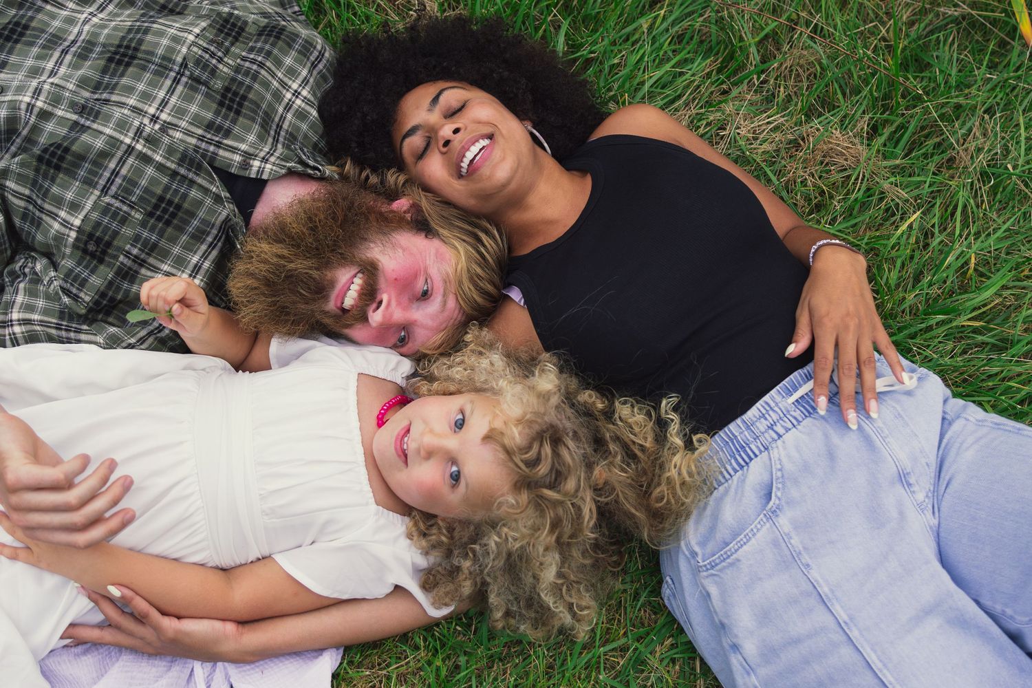 Three friends lying on green grass together, smiling and relaxing outdoors on a sunny day.