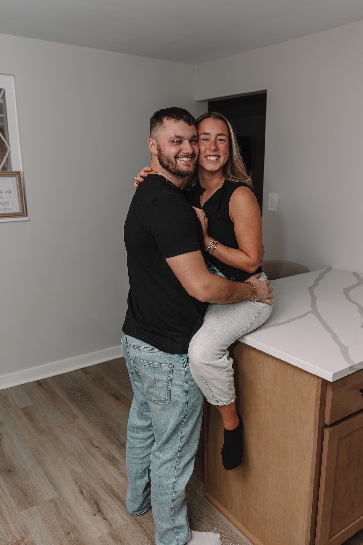 Happy couple embracing in a modern kitchen, woman sitting on counter, both smiling warmly in casual attire.