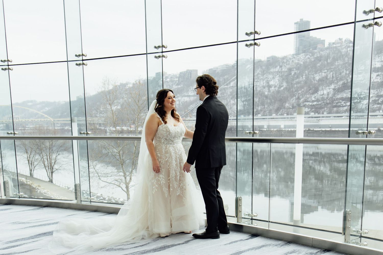 Bride and groom share a romantic moment by floor-to-ceiling windows overlooking a snowy river landscape at their wedding venue.