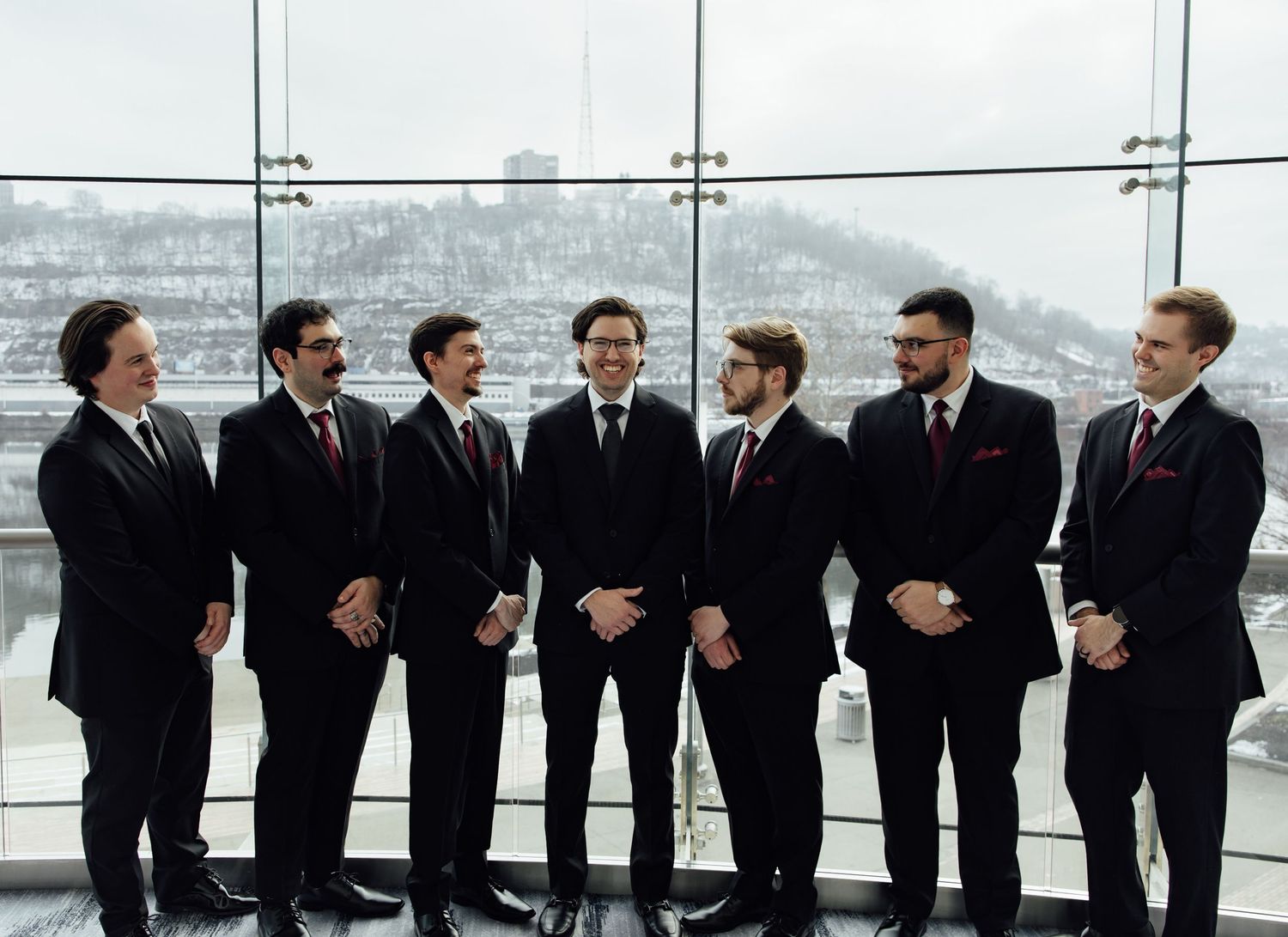 Seven men in black suits posing together indoors with a snowy cityscape visible through large windows.