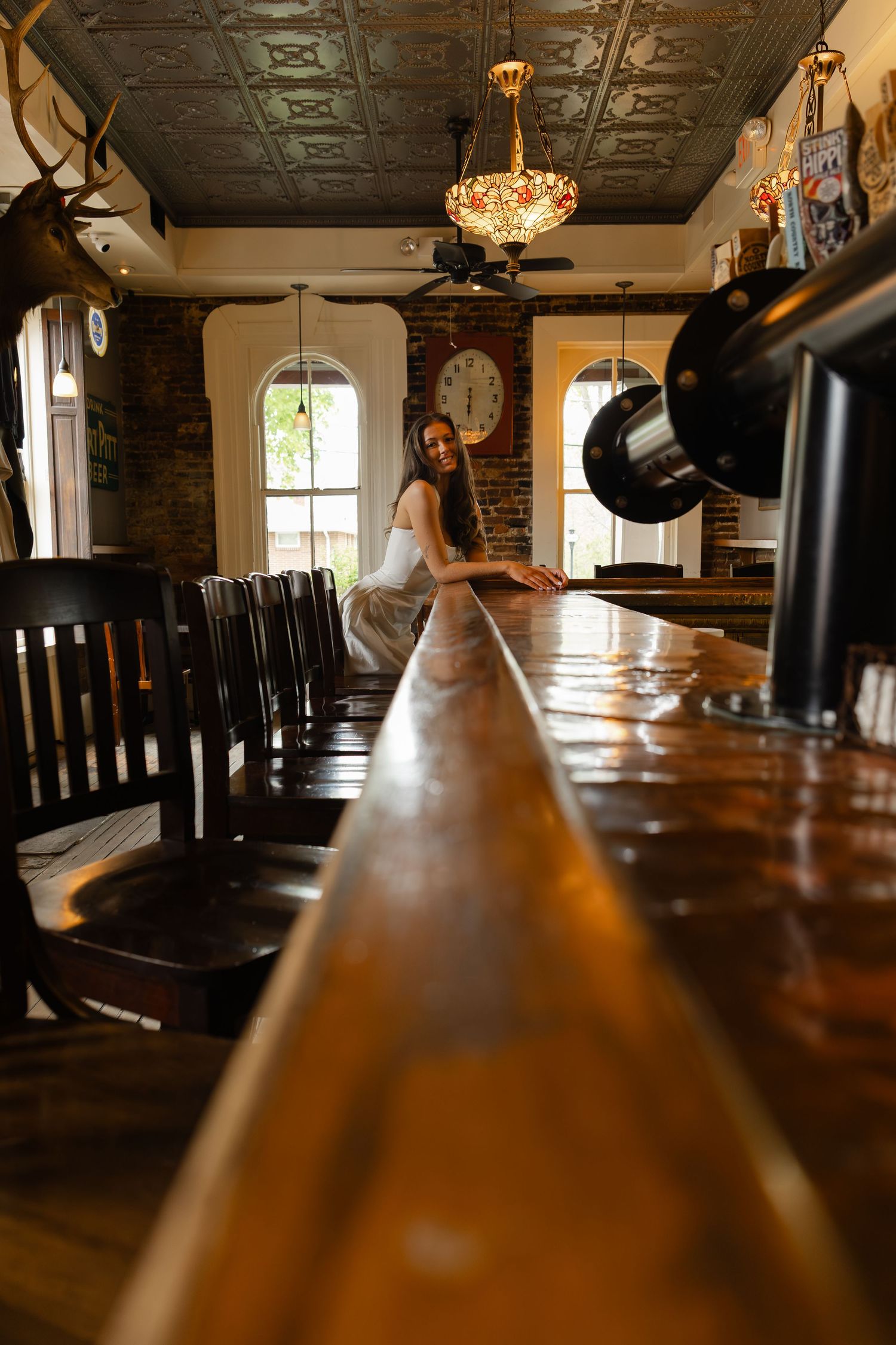Woman in white dress leaning on a long wooden bar in a vintage tavern with ornate ceiling and warm lighting.