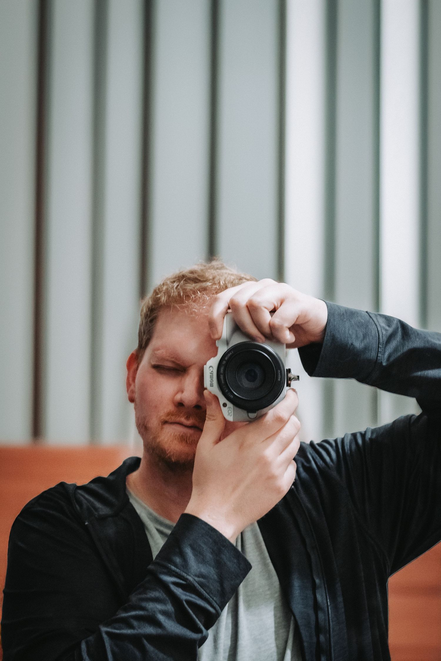 A red-haired man with a beard holds a mirrorless camera up to his face, photographing in a modern indoor setting.