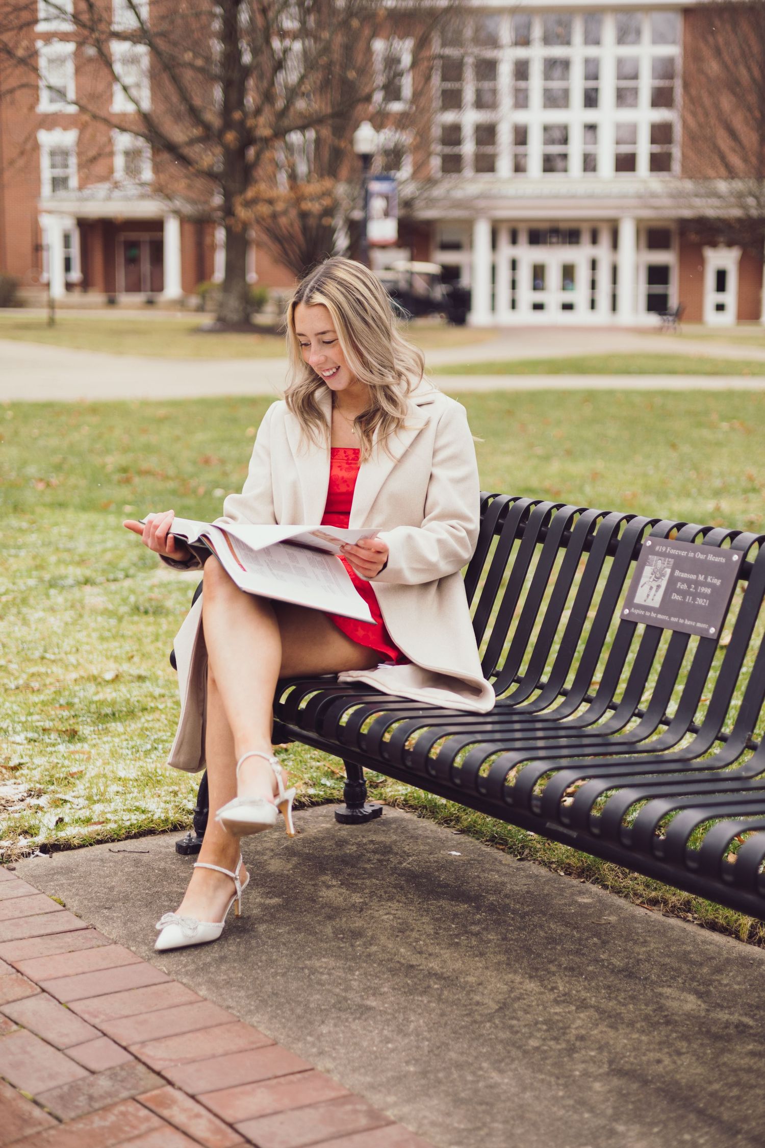 Graduate in light coat and red dress sits on metal bench reviewing papers outdoors.