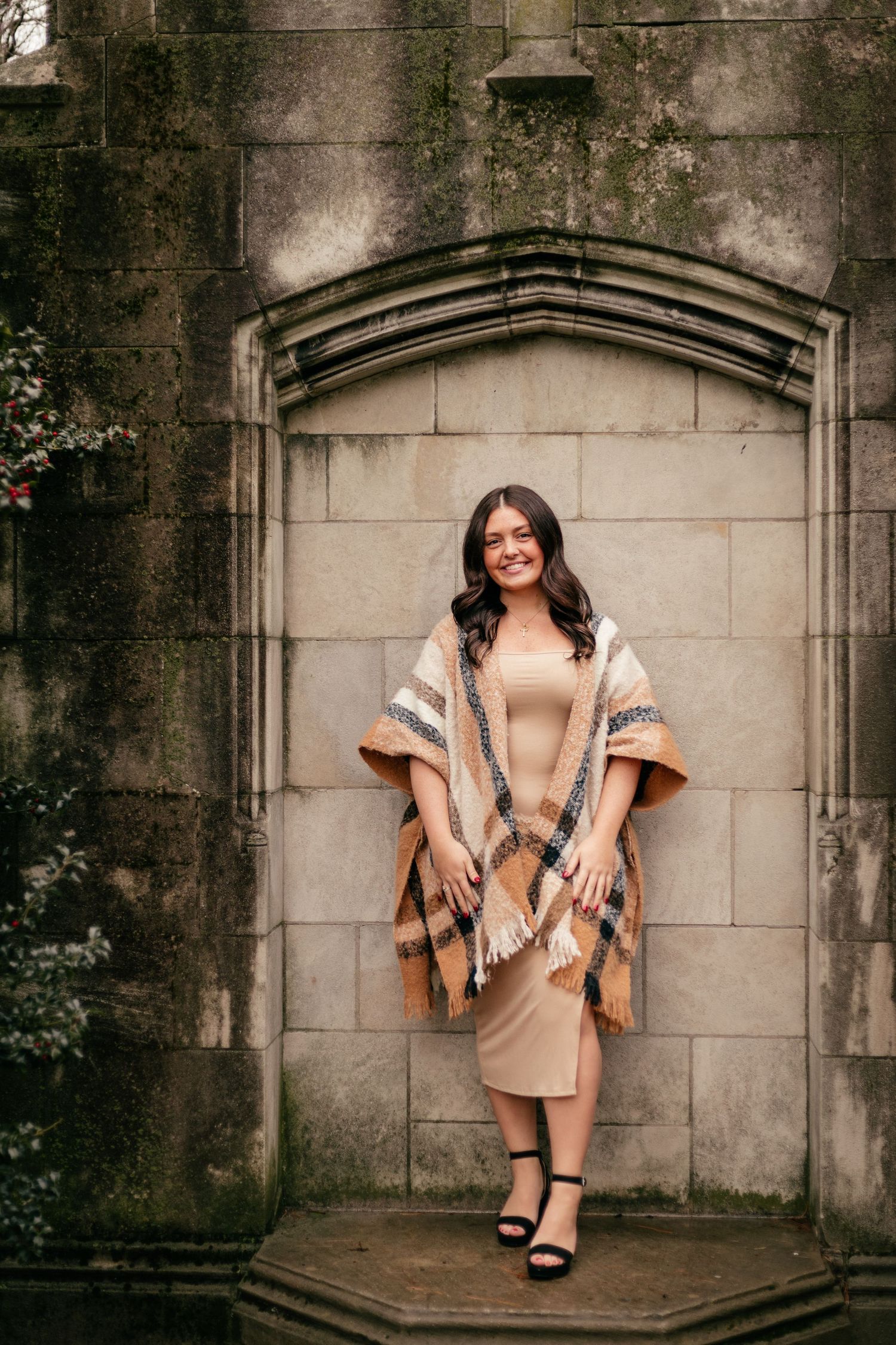 Woman in beige dress and striped poncho poses elegantly in stone architectural alcove.