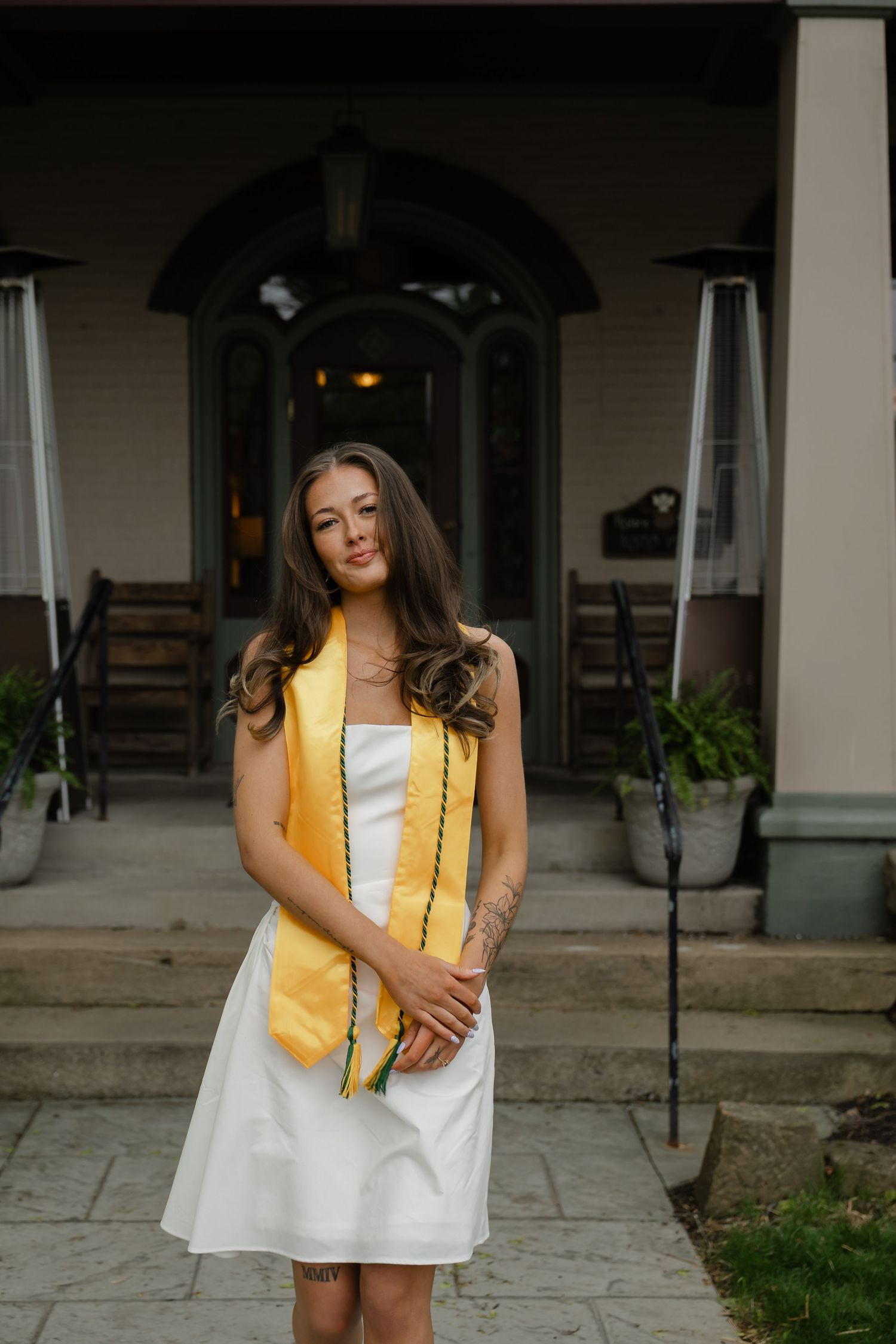 Young woman in white dress and gold graduation stole poses proudly in front of elegant building entrance.