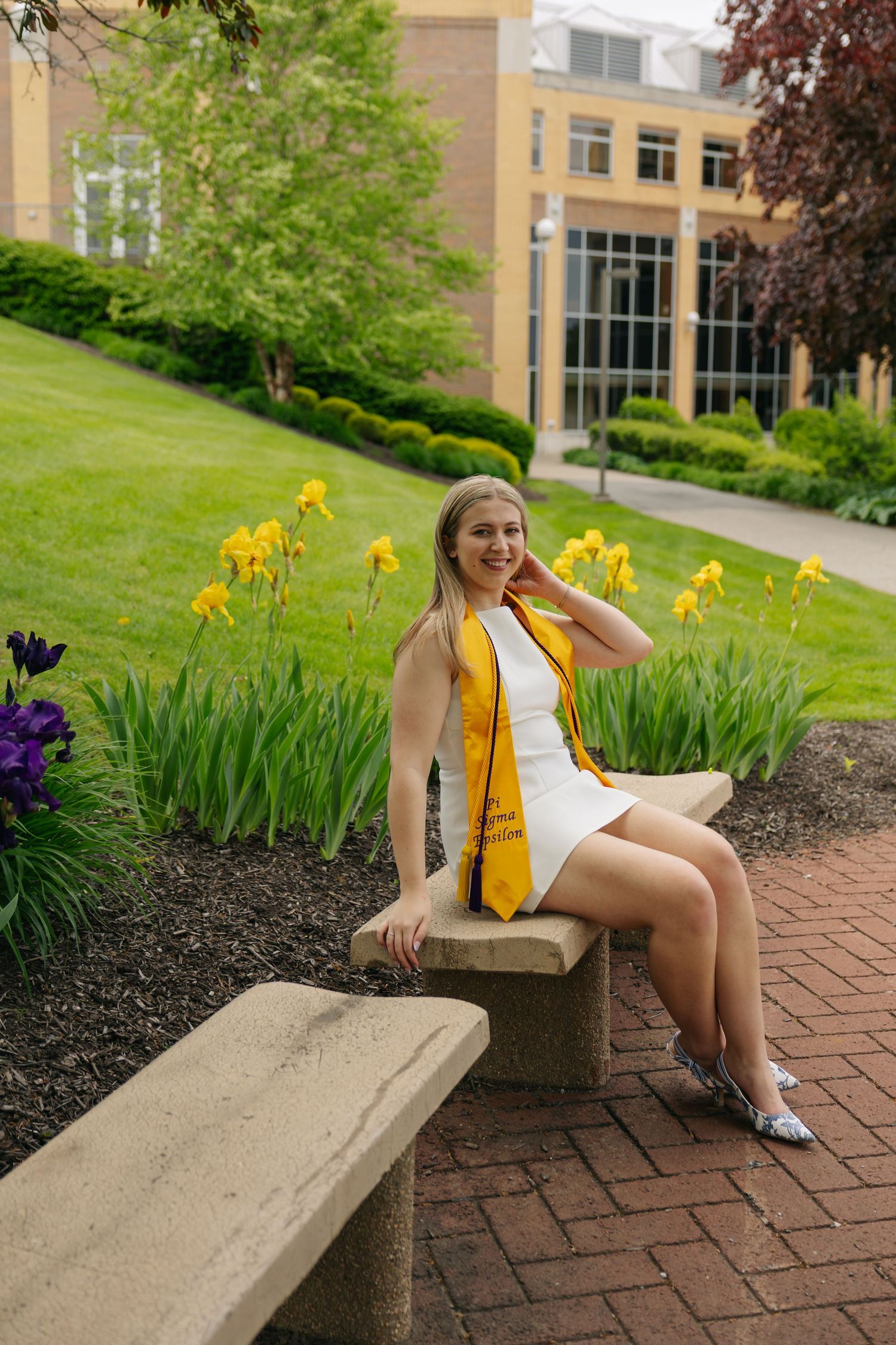 A smiling graduate in a white dress and gold stole sits on a stone bench outdoors near blooming flowers and a brick building.