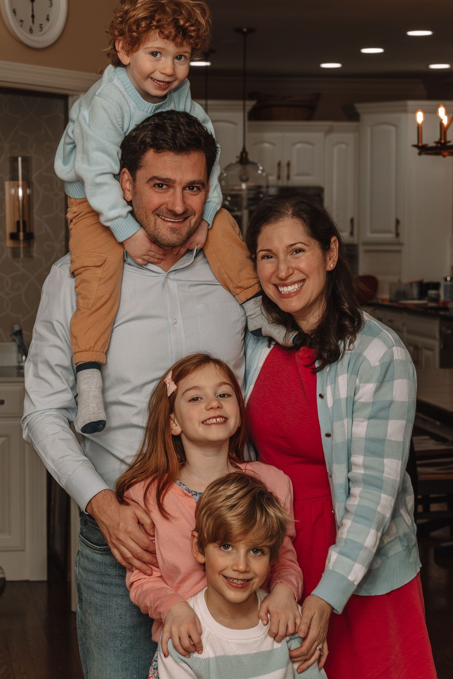 Happy family of five posing together in a cozy kitchen, with dad holding toddler on shoulders and mom in red dress.