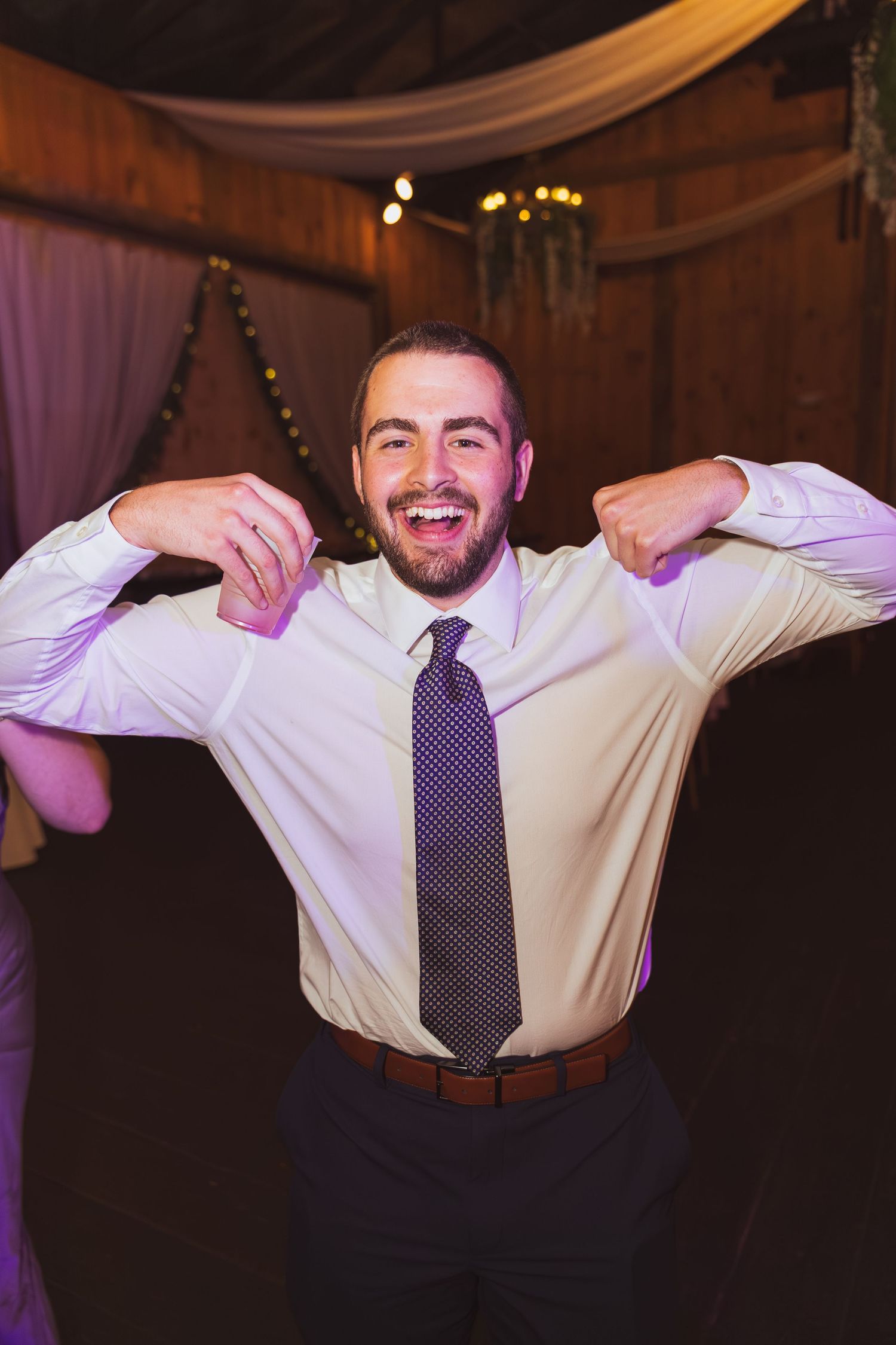 A smiling man in a white dress shirt and purple tie flexing his arms at a wedding reception.