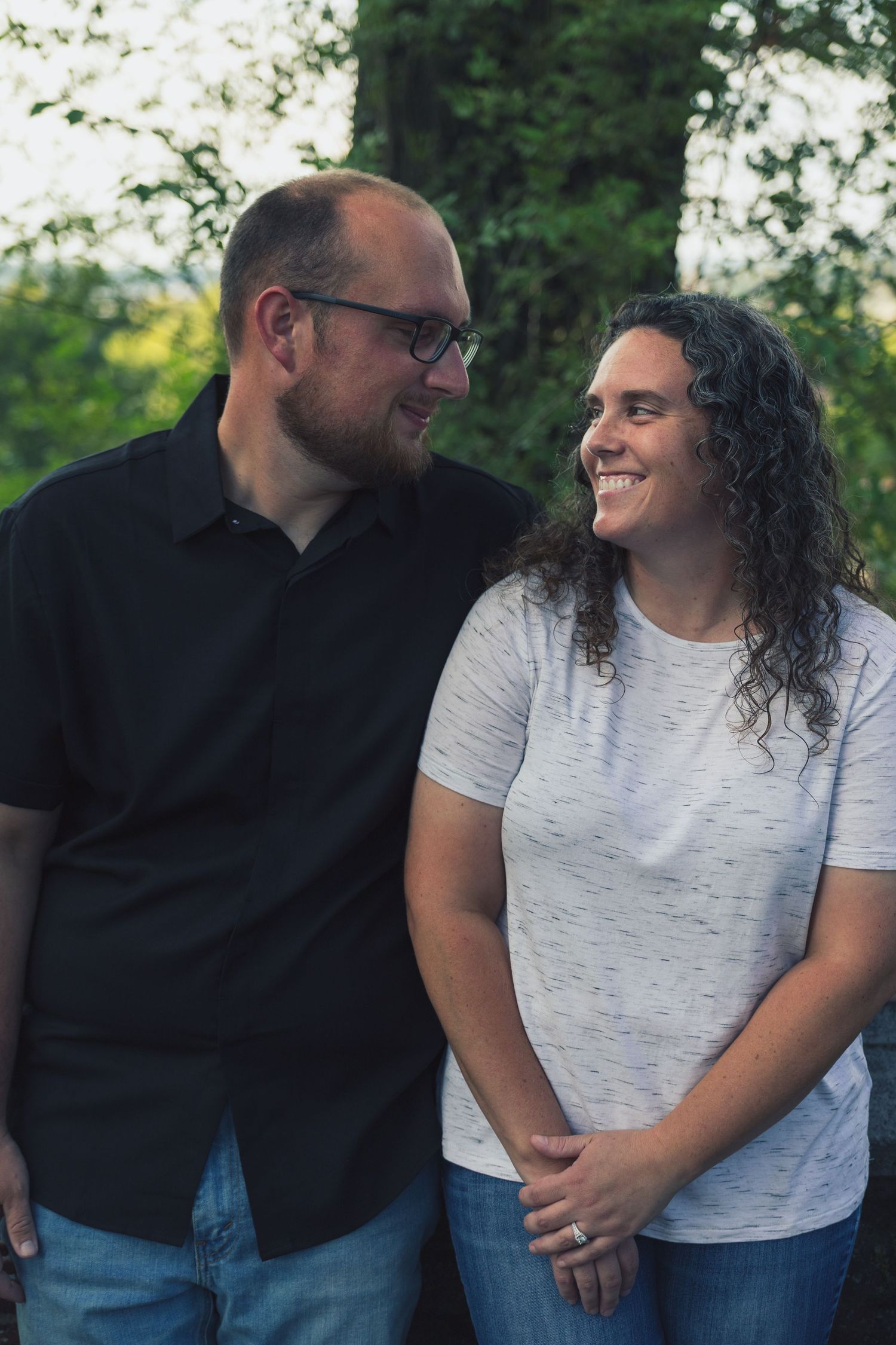 A smiling couple in casual attire sharing a tender moment outdoors with lush green trees in the background.