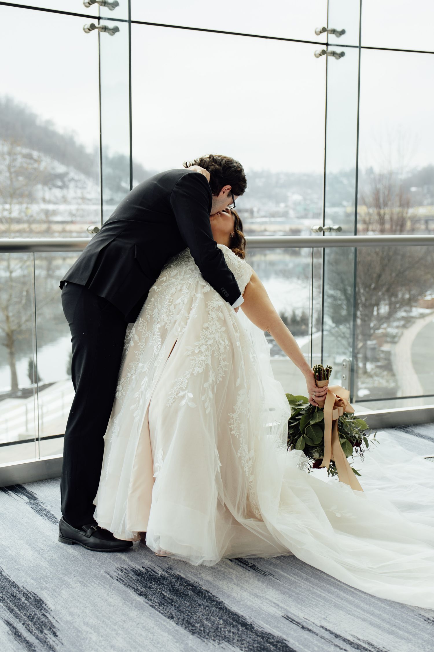 Bride and groom sharing a romantic kiss by a snowy winter window, bride holding a greenery bouquet in a lace ballgown.