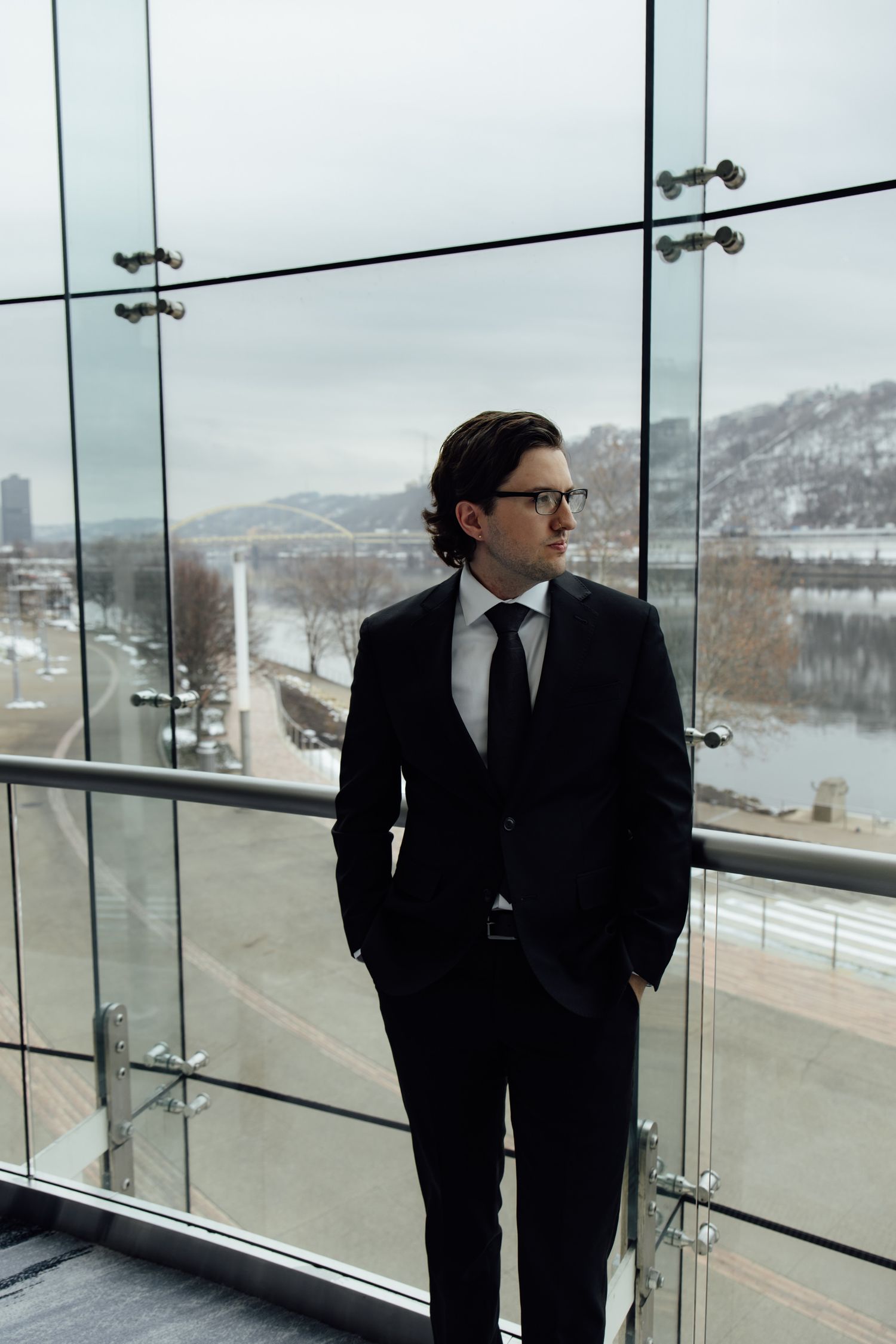 A professional man in a black suit and tie stands by a glass wall overlooking a snowy river cityscape.