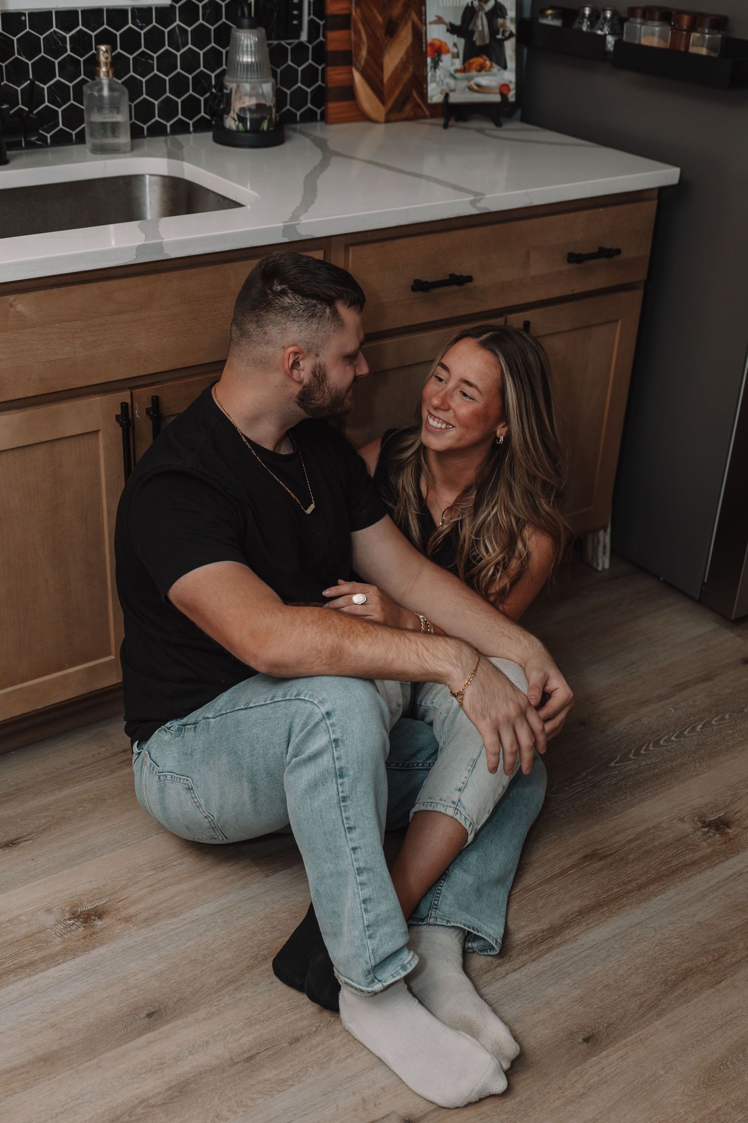 Happy couple sitting on kitchen floor laughing together, wearing casual jeans and black tops in modern home.