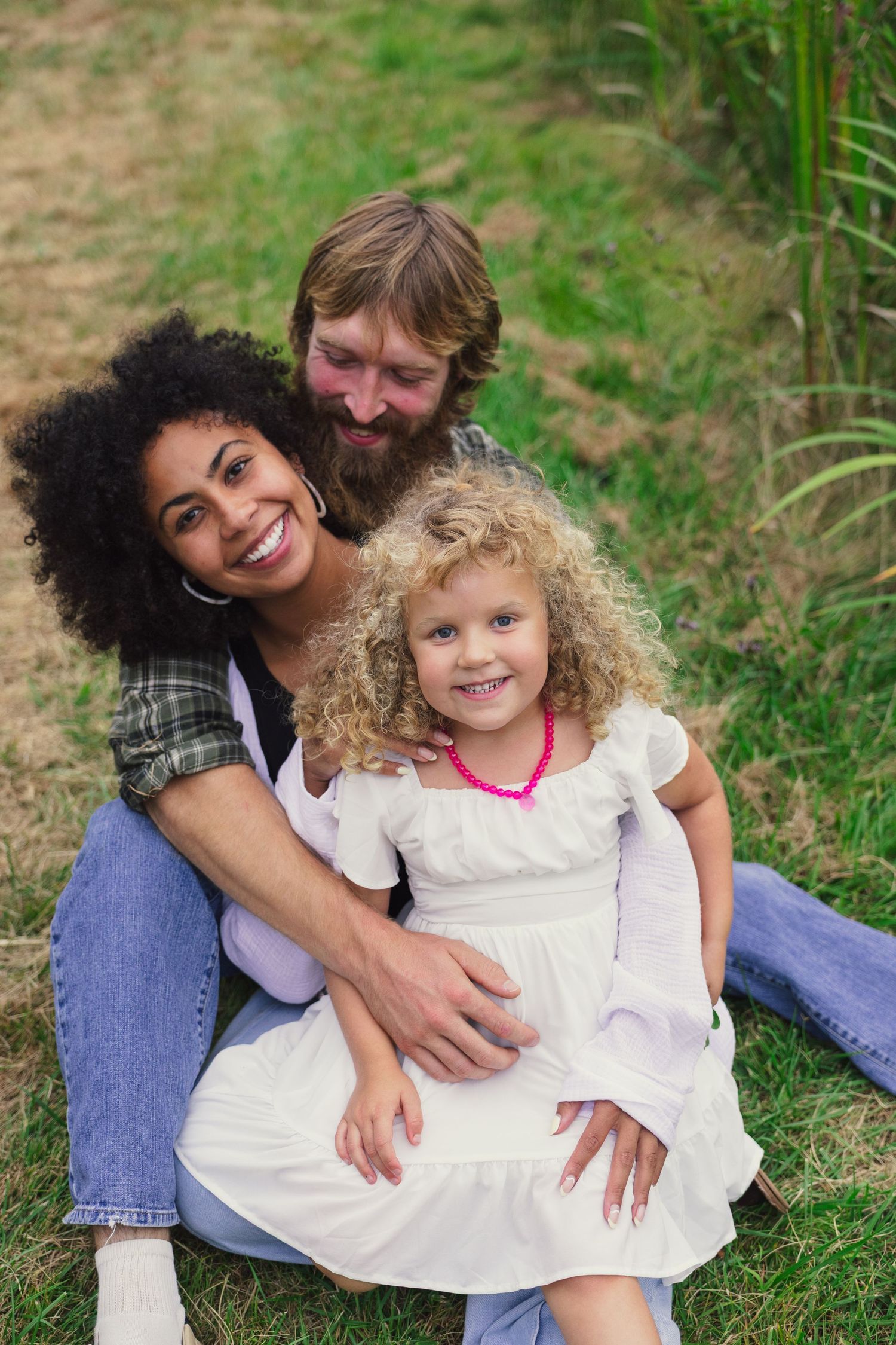 Happy interracial family sitting on grass, smiling mother, bearded father, and curly-haired girl in white dress outdoors.