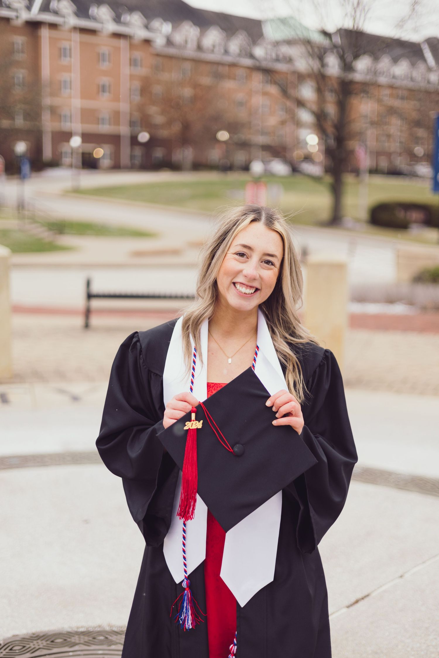 Graduate in black gown holding graduation cap while smiling on campus.
