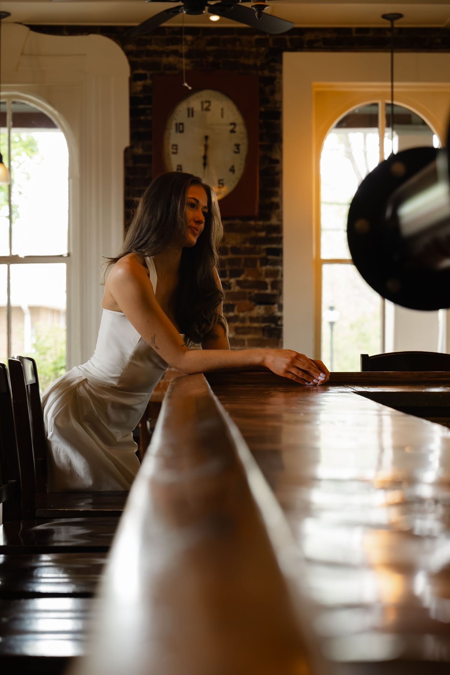 Young woman in white dress leaning on a long wooden bar in a rustic brick interior.