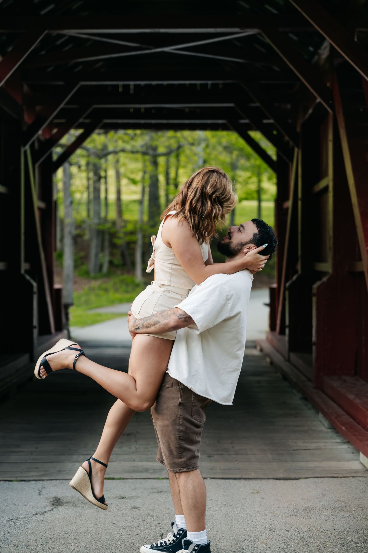 A man lifts a woman for a romantic kiss inside a rustic covered bridge surrounded by lush green trees.