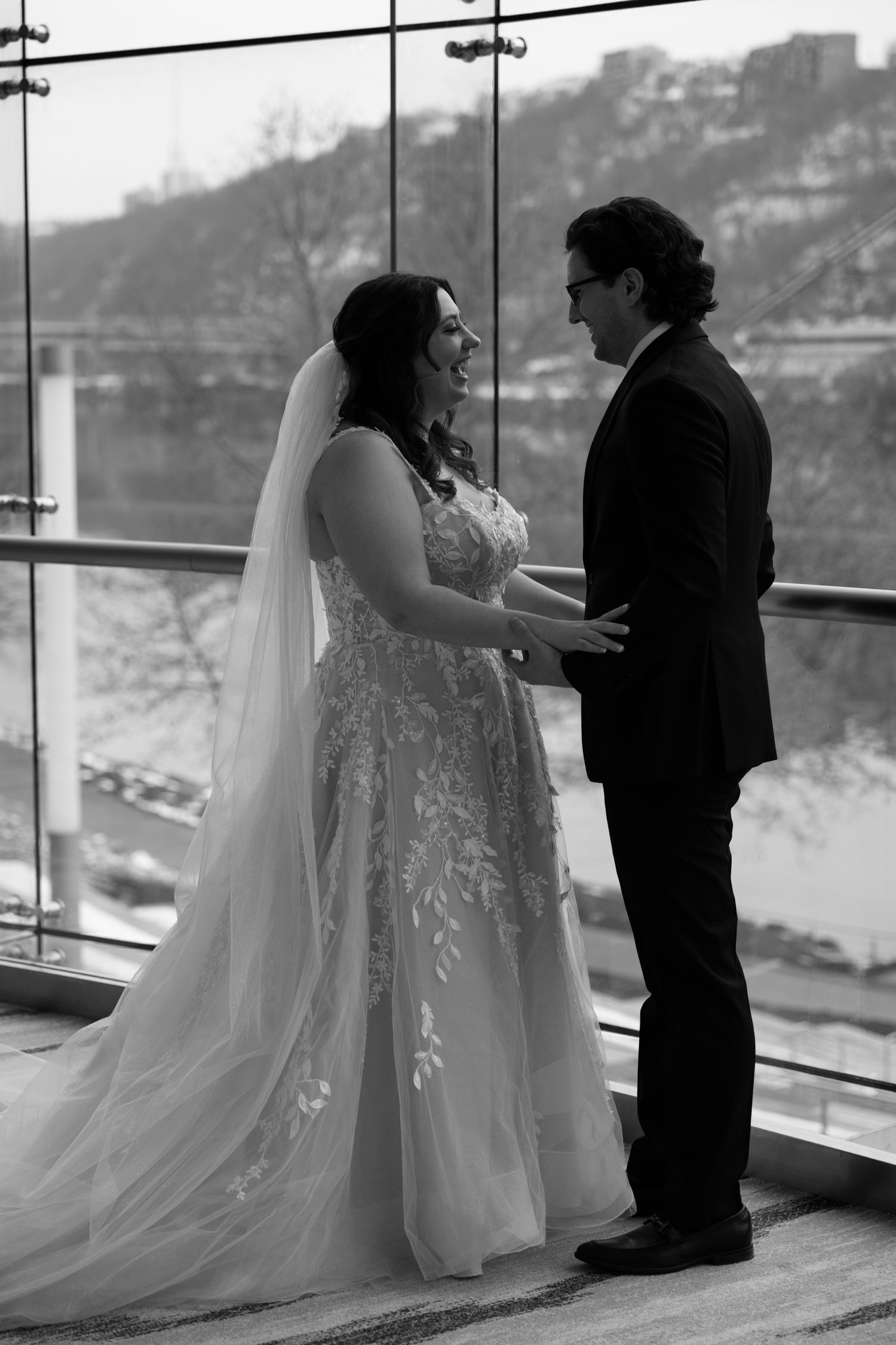 Black and white photo of a bride and groom holding hands, sharing a tender moment by a large window on their wedding day.
